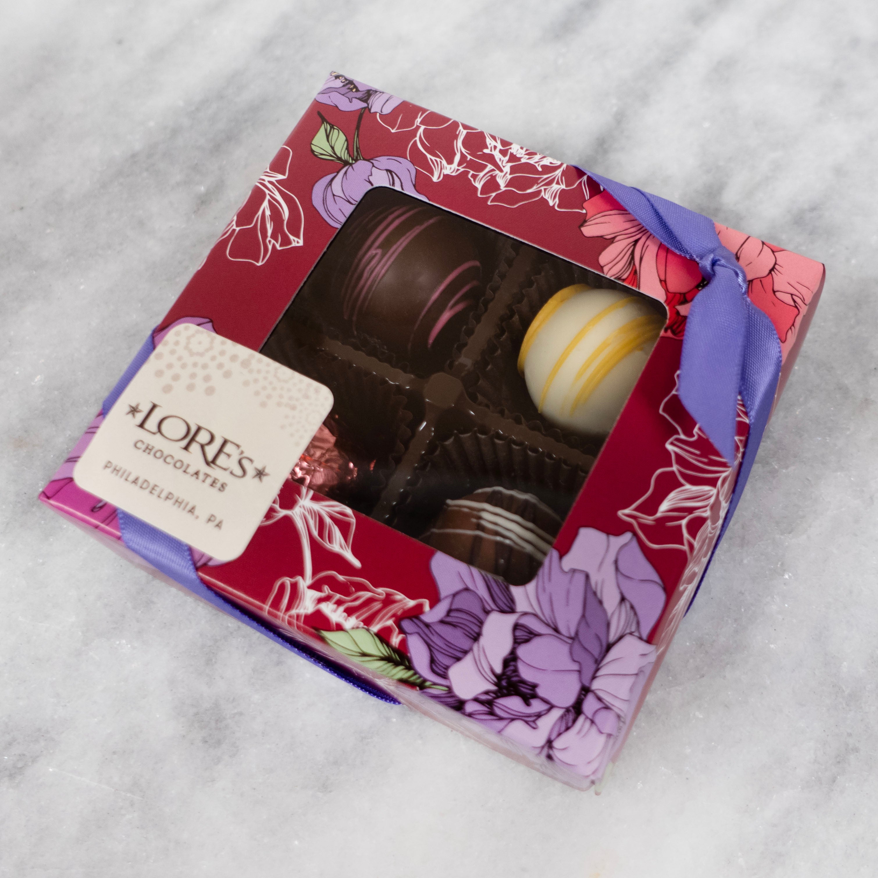 4 piece truffle assortment - design is a dark magenta with purple and white flowers and a clear window middle, sticker reads lore's chocolates philadelphia pa