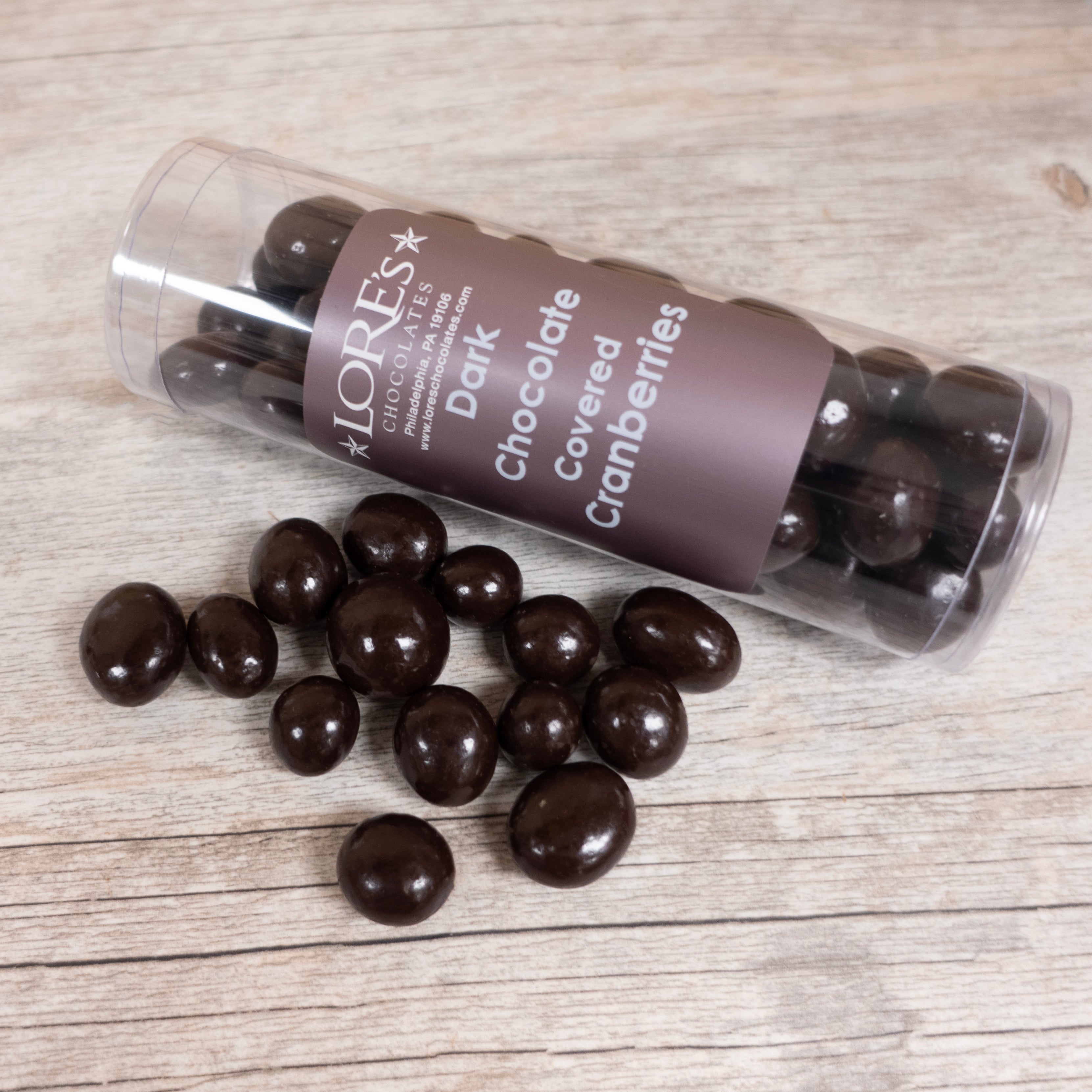 Dark chocolate-covered cranberries spilling out of a cylindrical container labeled 'Lore's' on a wooden surface.