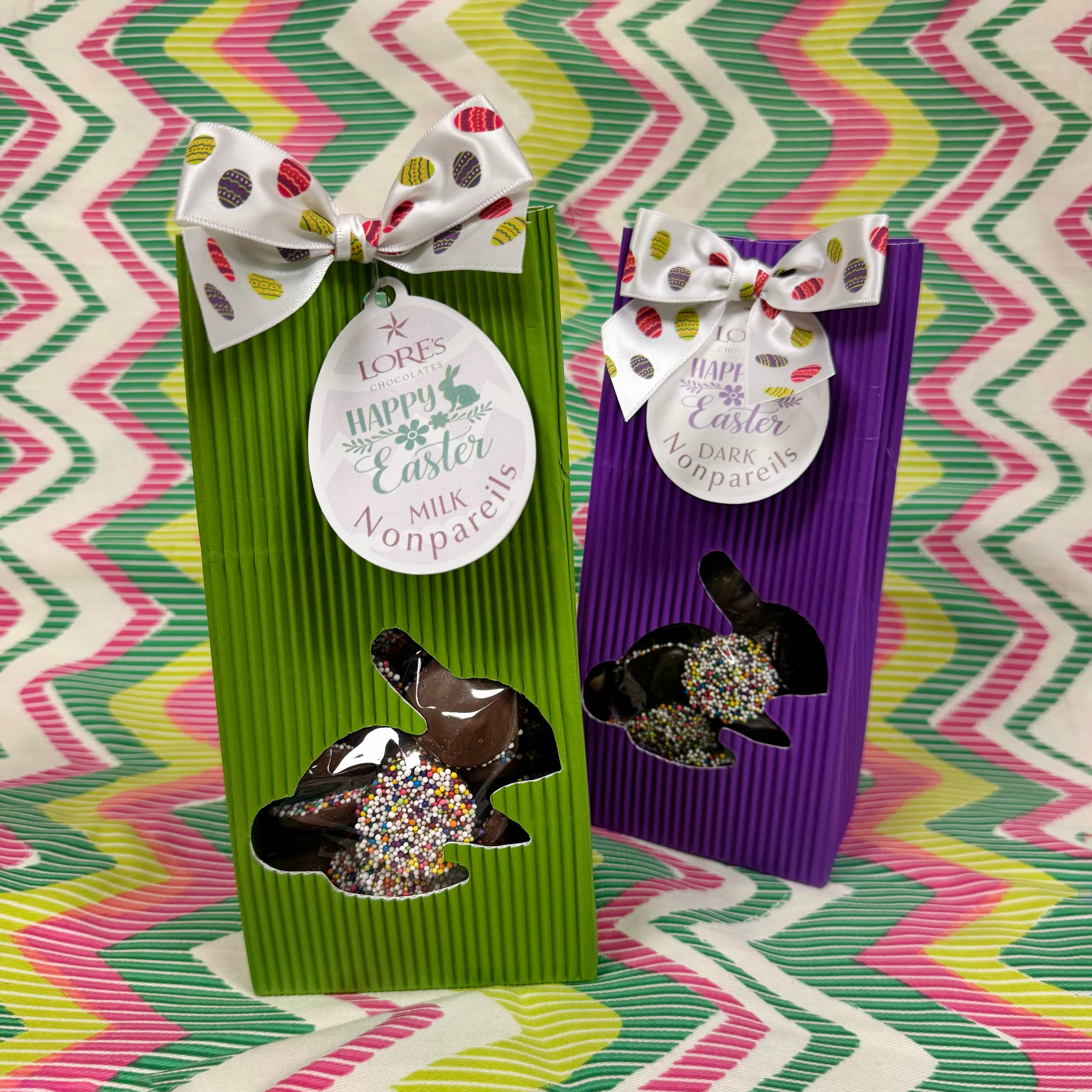 Two corrugated boxes of chocolate nonpariels in green and purple packaging with Easter-themed bows on a colorful zigzag background.