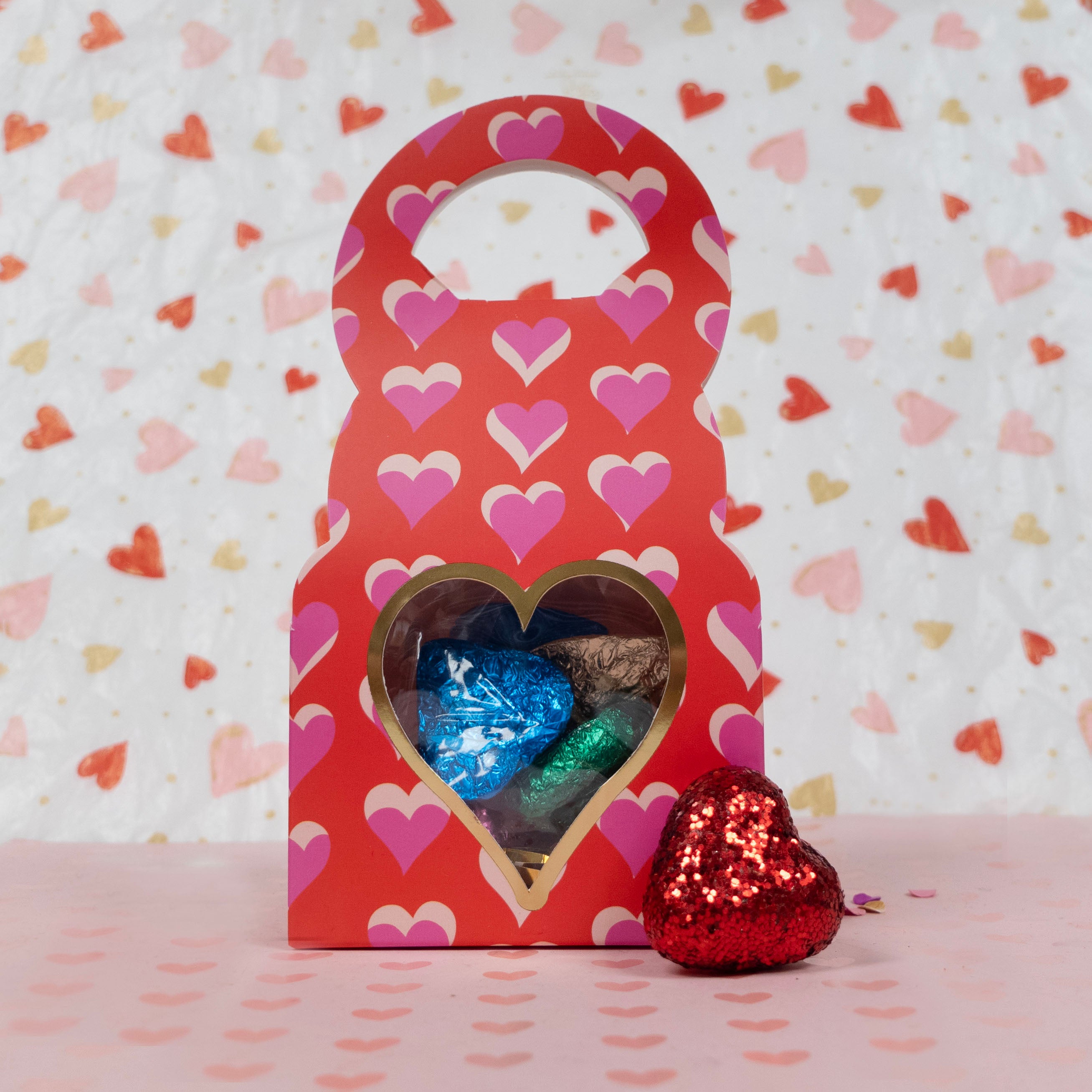Red gift box with heart-shaped window containing foil-wrapped chocolates on a heart-patterned background.