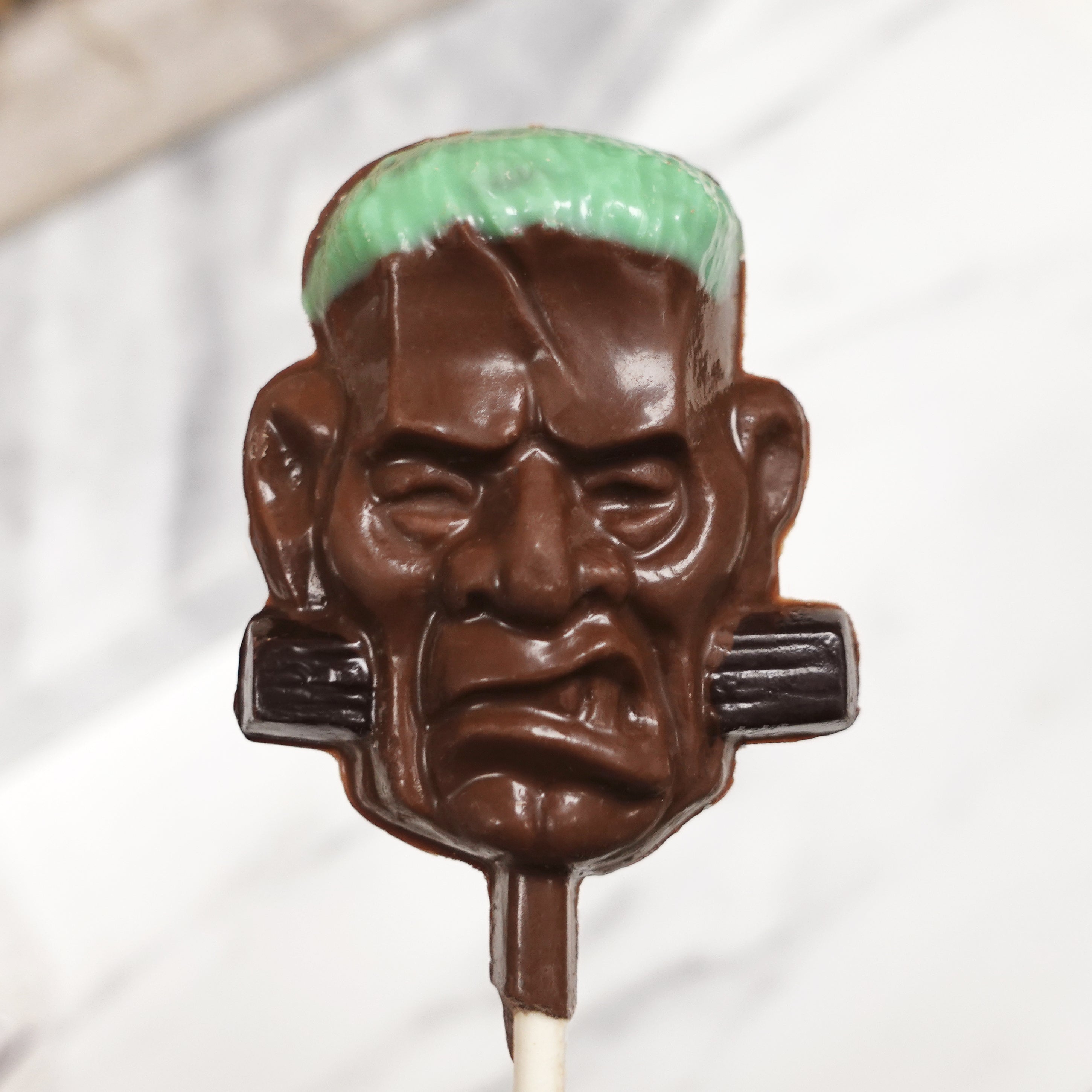 Chocolate lollipop shaped like a Frankenstein's monster face with a green top on a white background