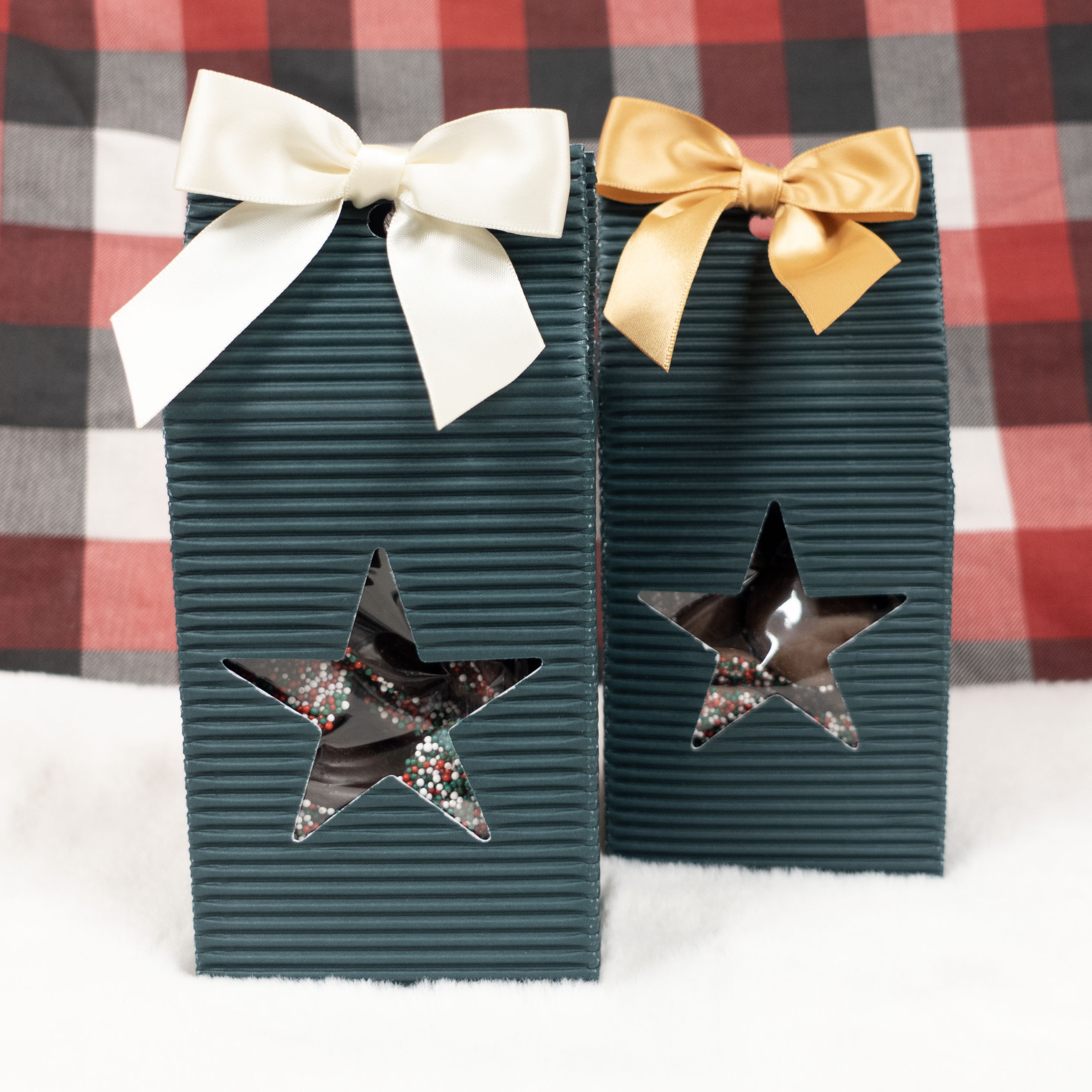 Two green boxes of Christmas nonpareils with white and gold ribbons with star cutouts on a holiday checkered background