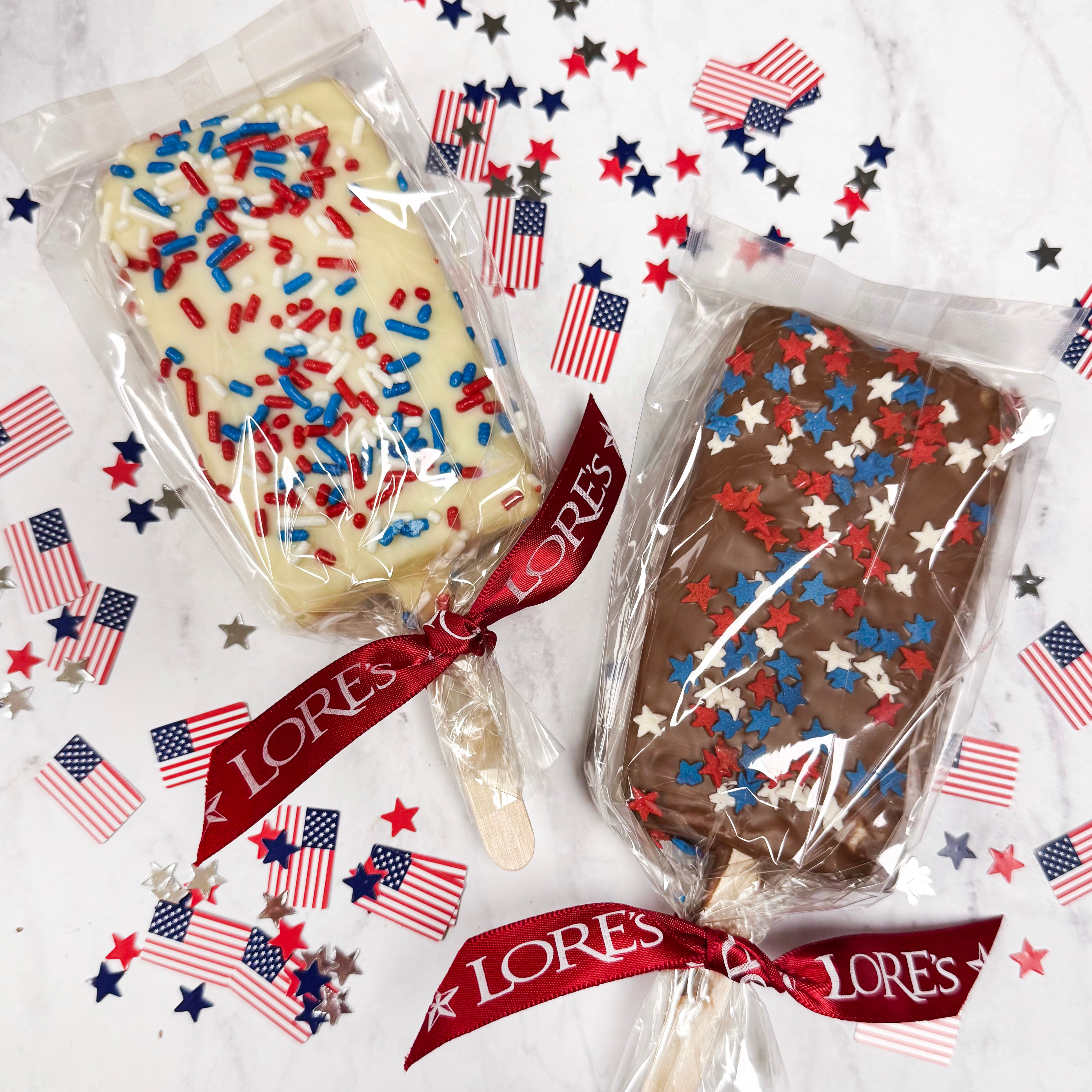 Two rice crispy lollipops covered with milk or white chocolate with patriotic designs, sprinkles, and jimmies, and 'LORES' branding on a decorative background. Lore's Chocolates in Philadelphia