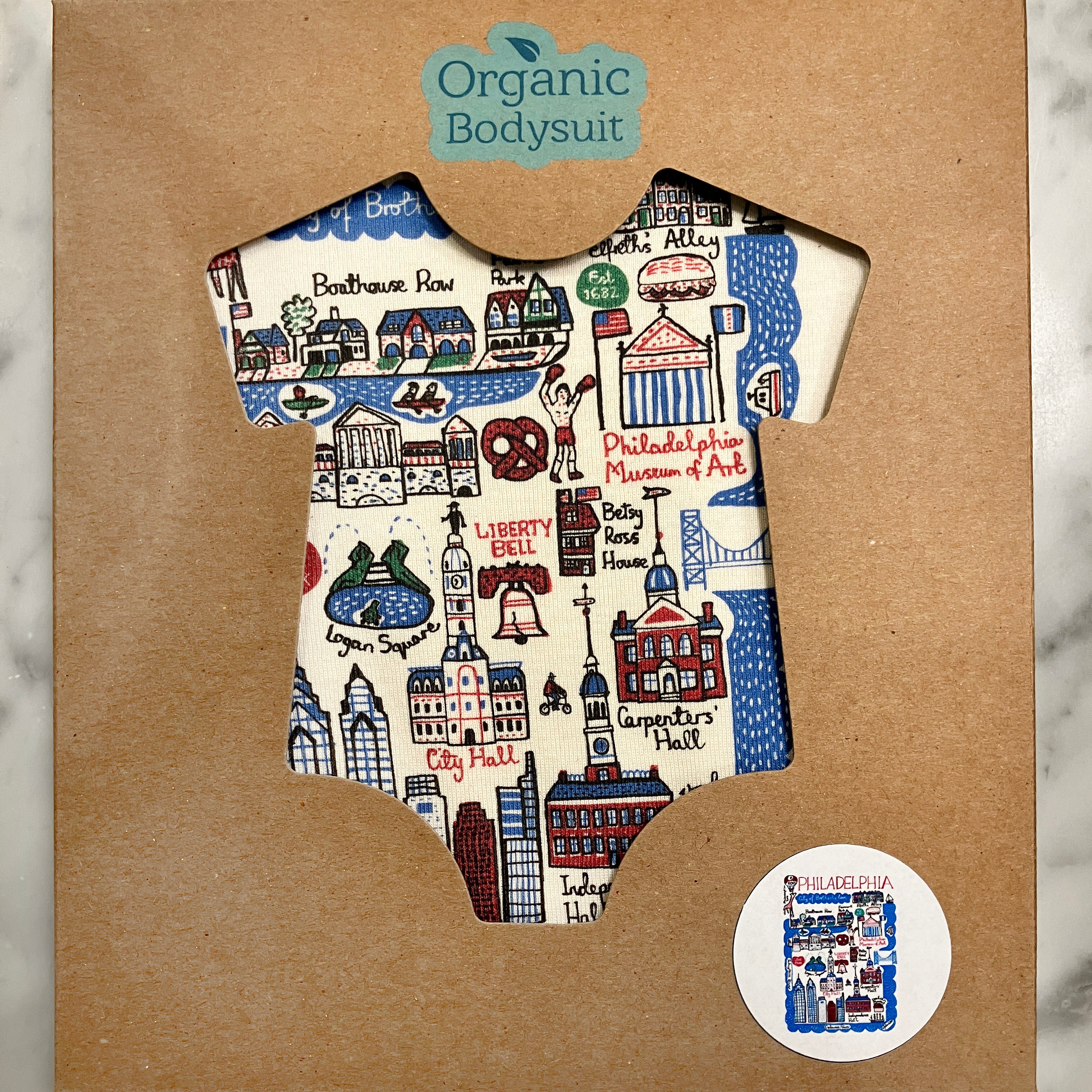 Organic baby bodysuit with Philadelphia cityscape design in a cardboard cutout of a bodysuit, branded 'Organic Bodysuit'.