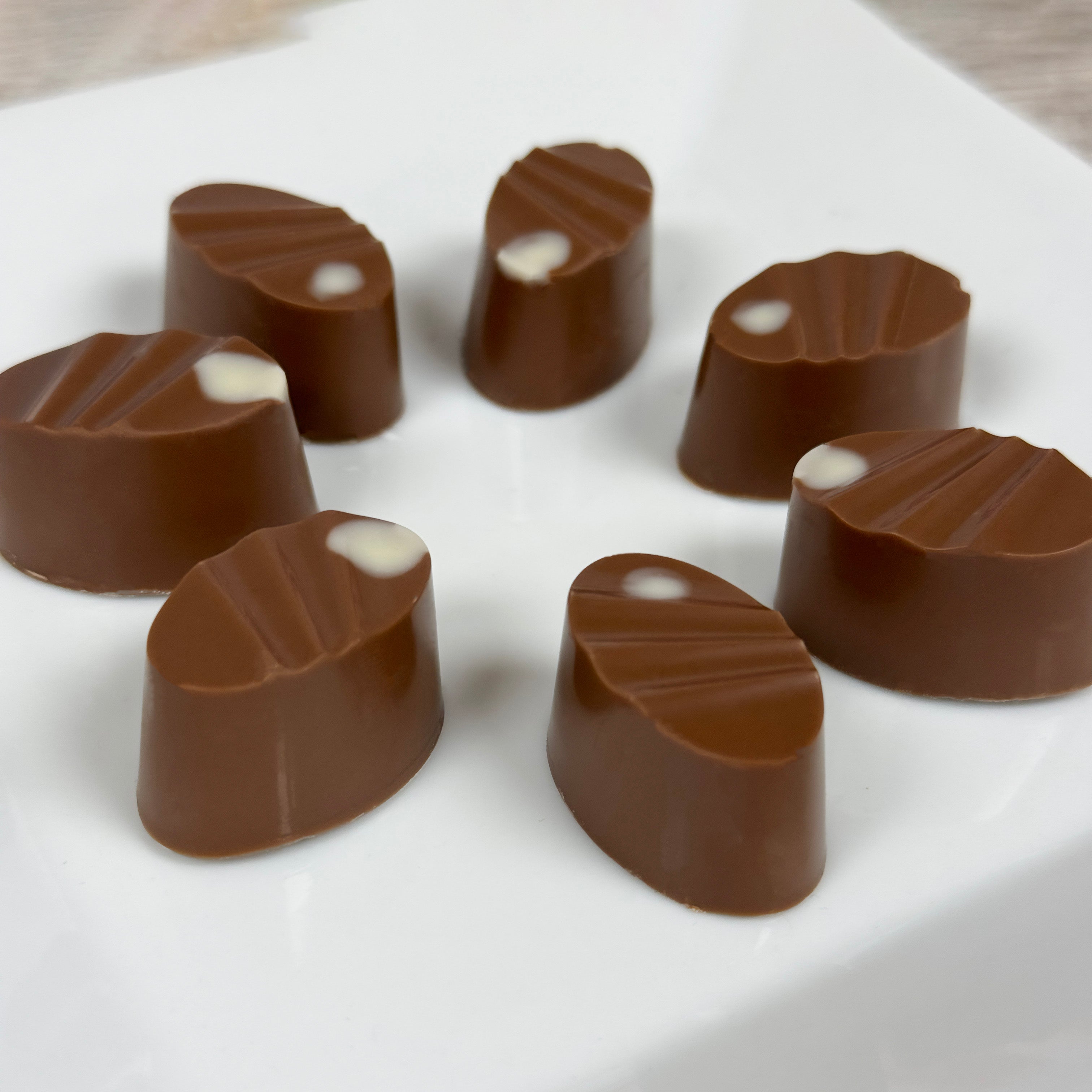 Six oval chocolate caramels arranged in a circle on a white square plate. Each truffle is brown with a small white inlay near the top.