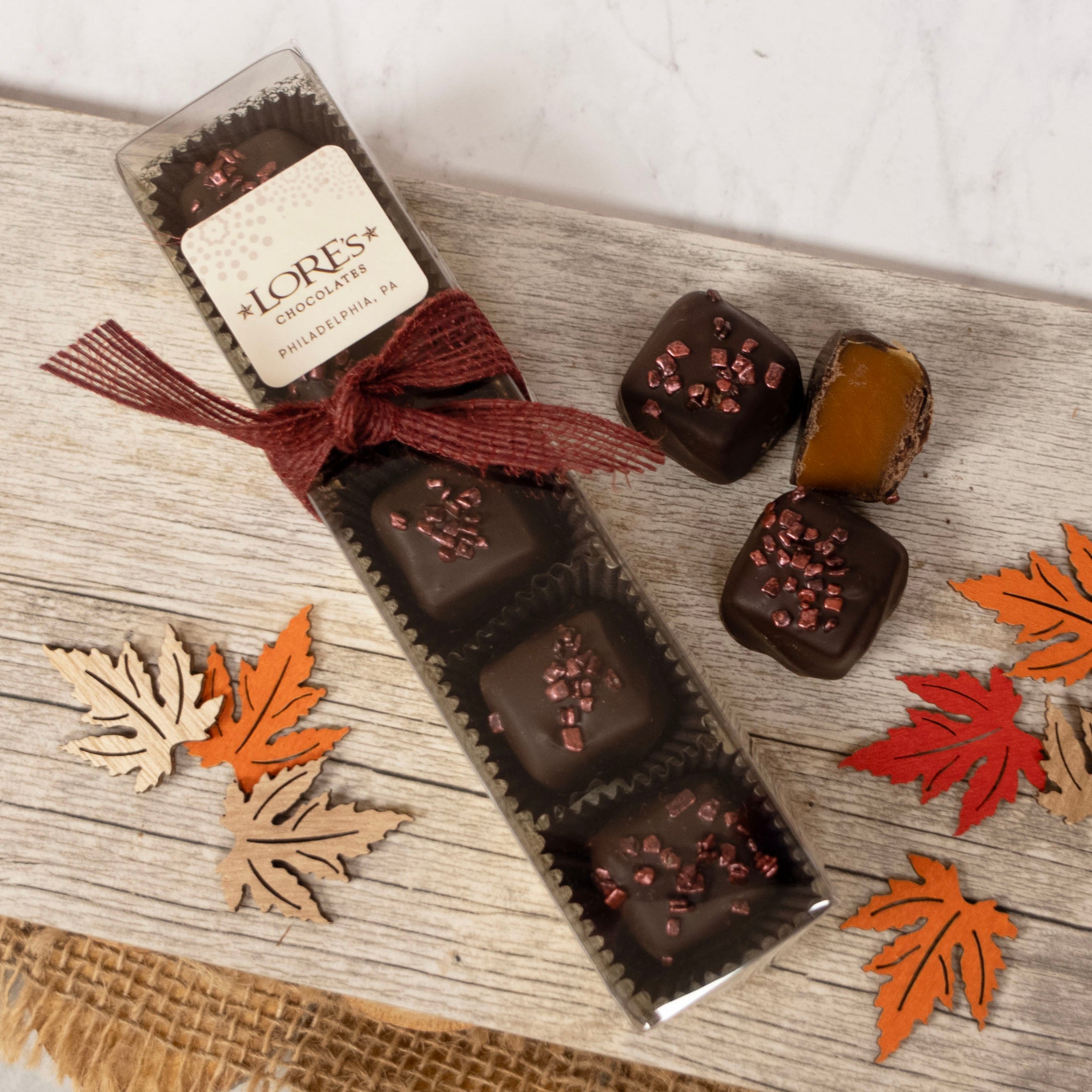 Box of chocolates (dark chocolate caramels) with a bow on a wooden surface with autumn leaves.