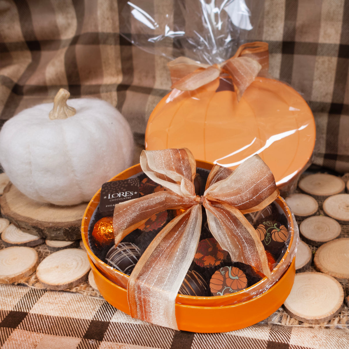 An orange gift box with a 'Pumpkin' theme, containing chocolate truffles, adorned with pumpkin and leaf designs. The box is placed on a textured surface, with a 'Burberry' checkered pattern in the background and a pumpkin to the left.