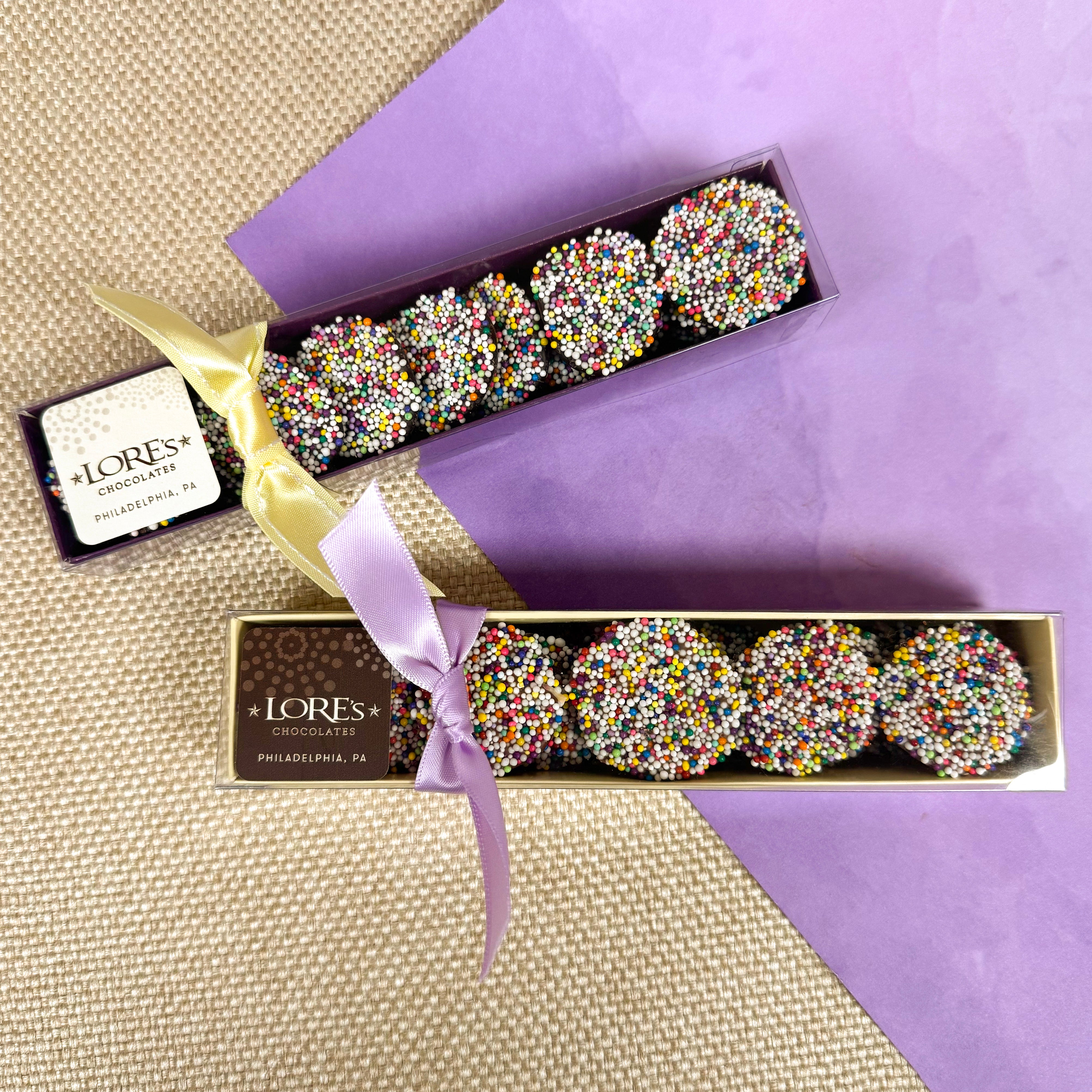 Two boxes of colorful chocolate nonpareils with a gold ribbon on a textured surface.