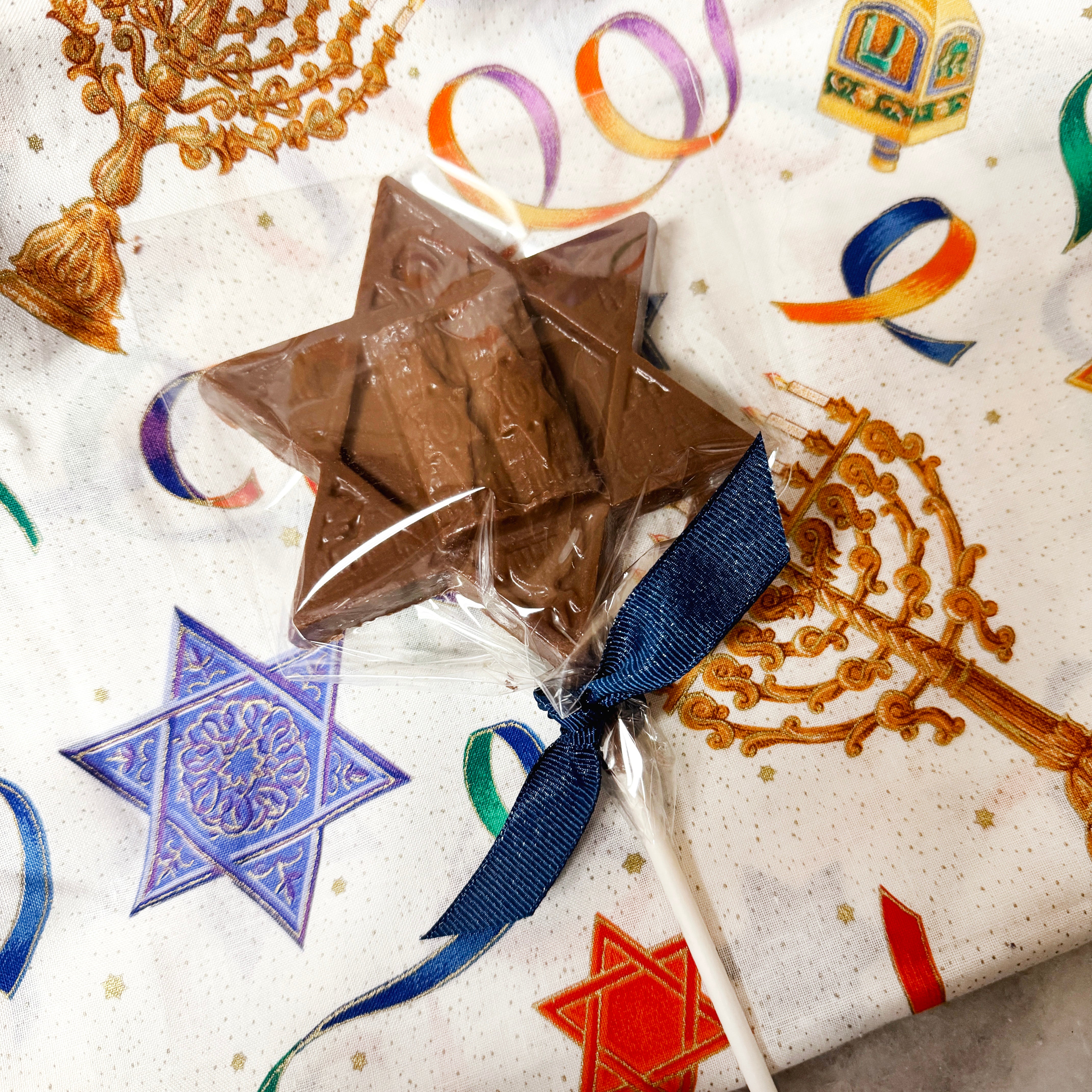 A chocolate lollipop with a Star of David shape, wrapped in a clear wrapper with a blue ribbon, displayed against a background with Hanukkah-themed patterns.
