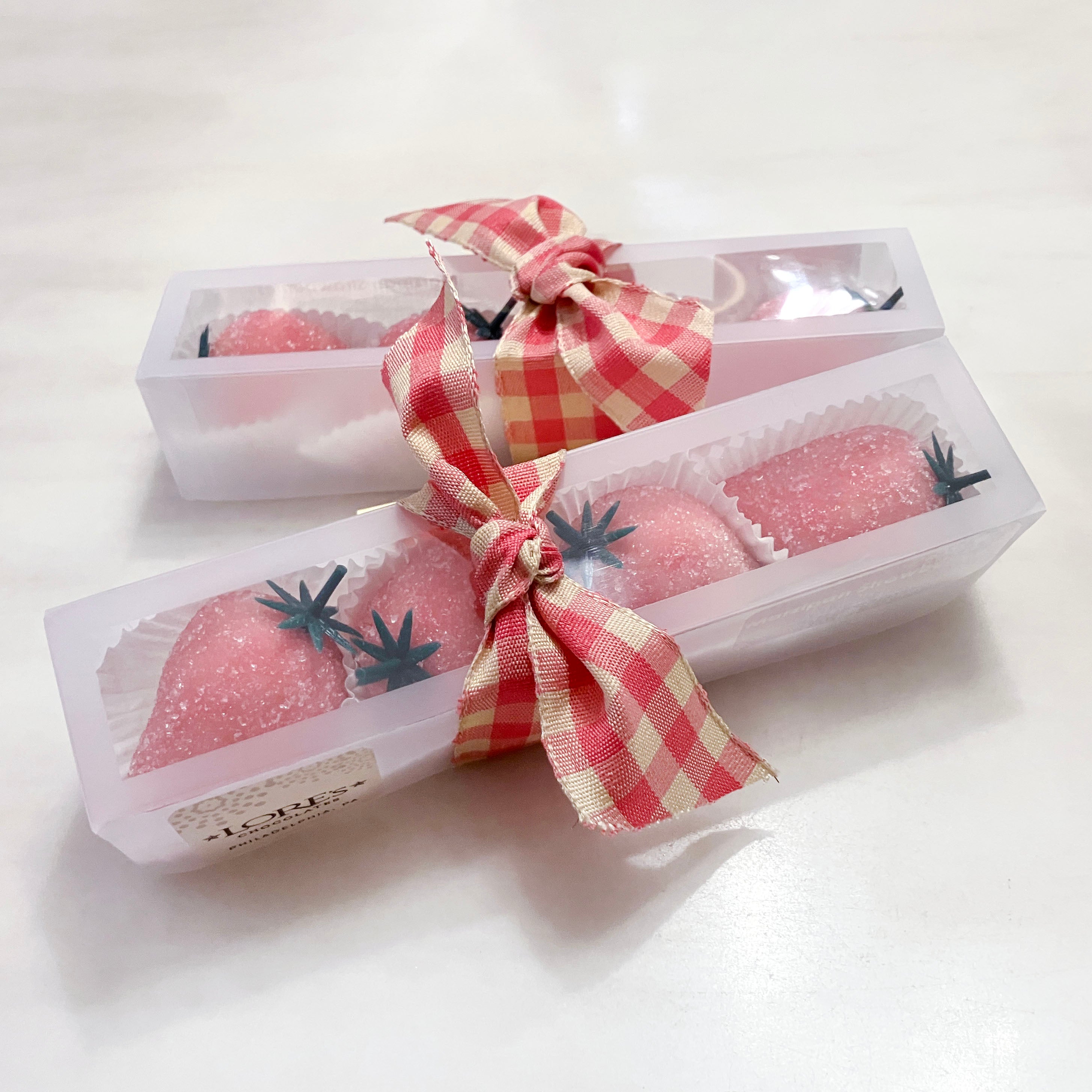 Pink strawberry shaped treats in clear boxes with red and white checkered ribbons on a white background