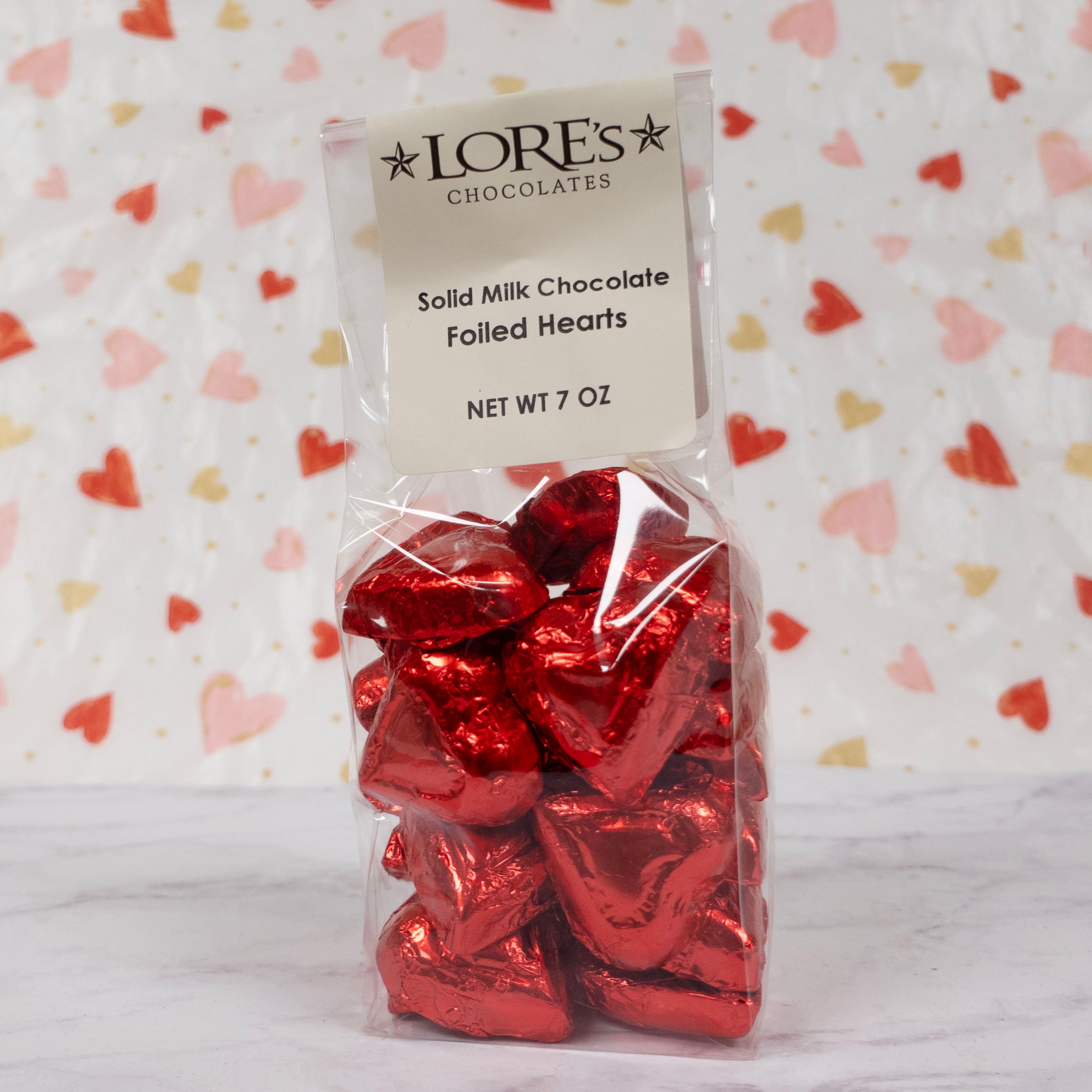 Pack of red foil-covered chocolates in heart-shapes with LORES Chocolates packaging on a heart-patterned background