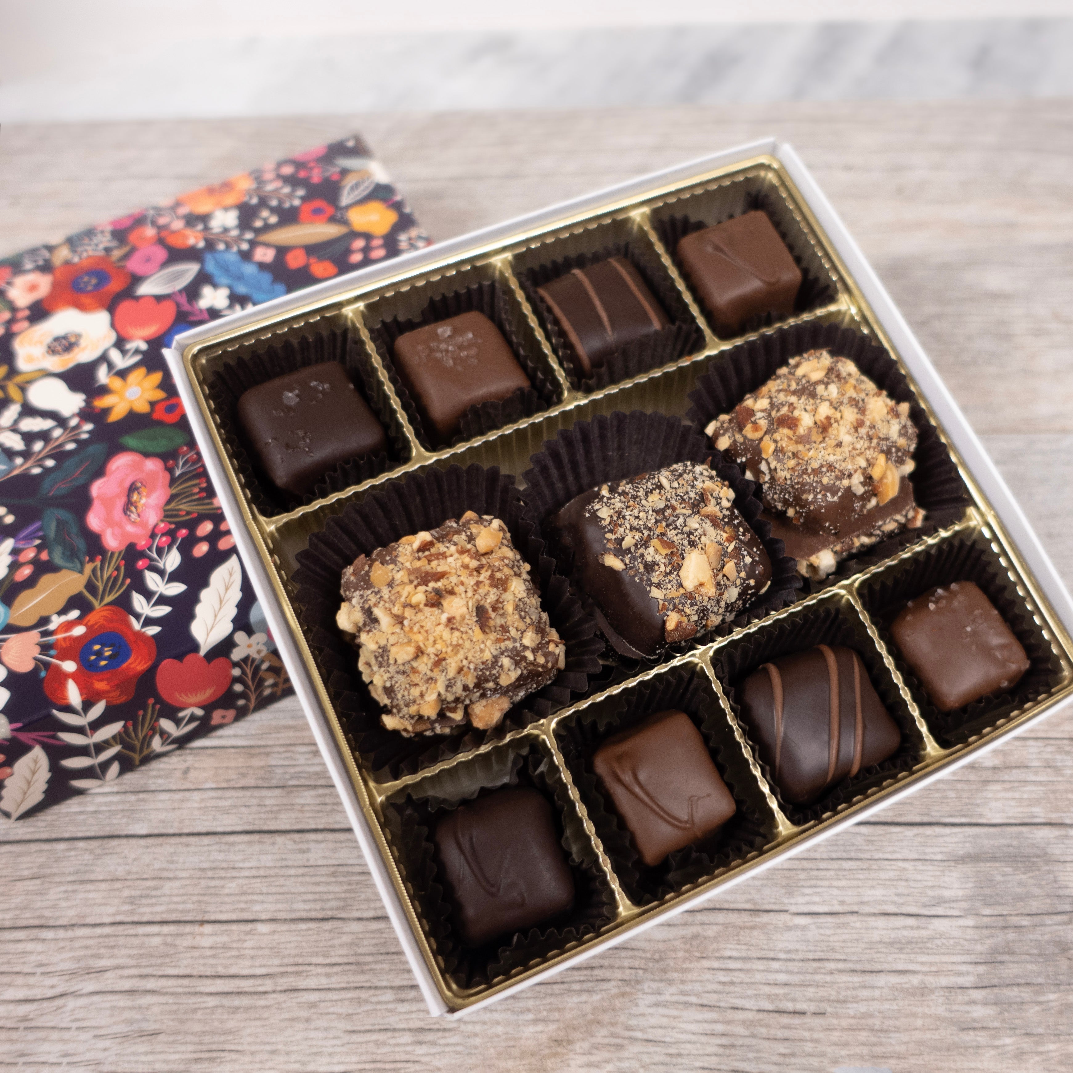 Assorted chocolates in a box on a wooden surface with a floral cloth.