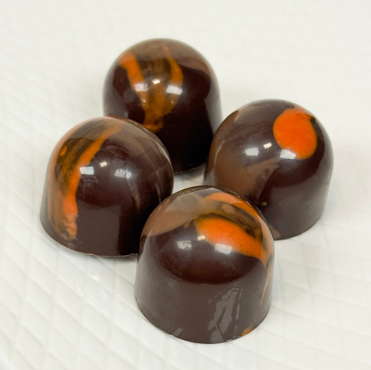 Four glossy dark chocolate domed confections with orange swirls, arranged on a white textured surface.