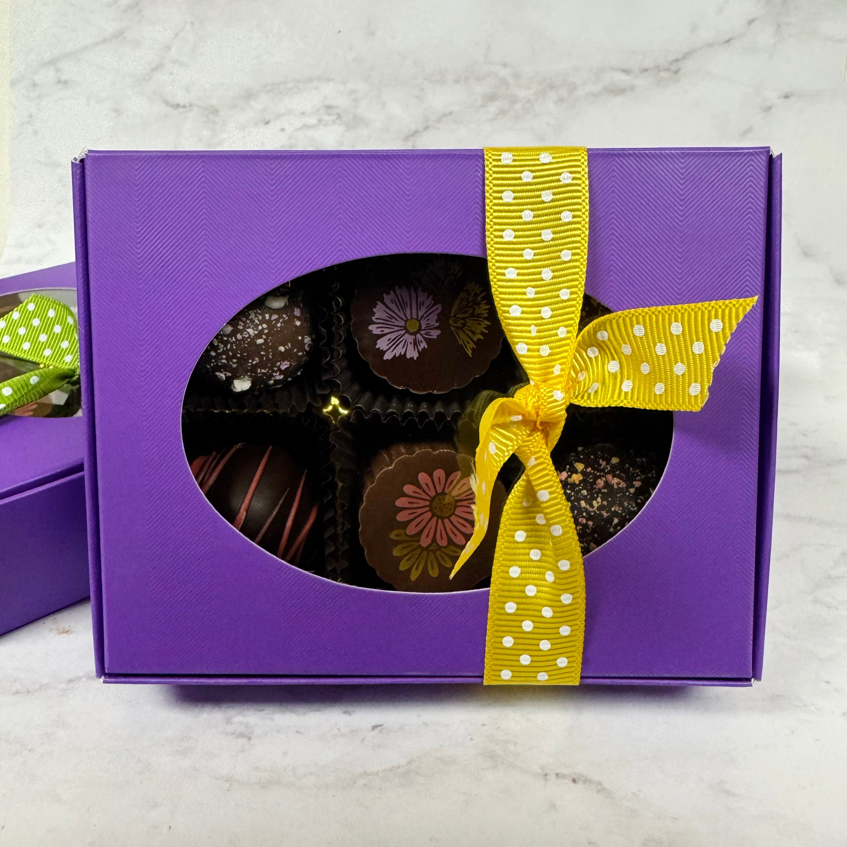 Purple gift box with a window, filled with chocolates, and a yellow ribbon on a marble surface.