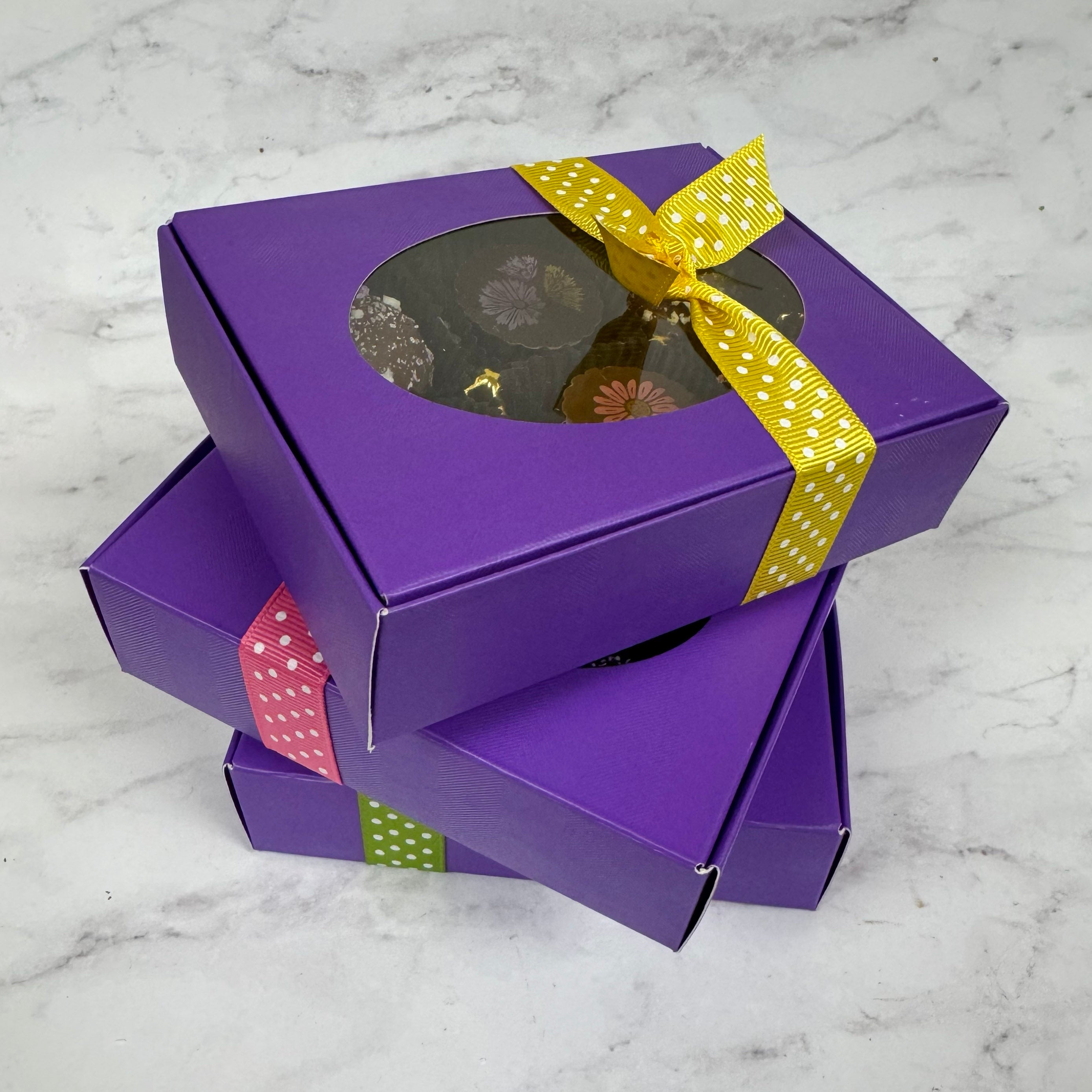 Purple gift boxes of chocolate truffles with a window and colorful ribbons on a marble surface.