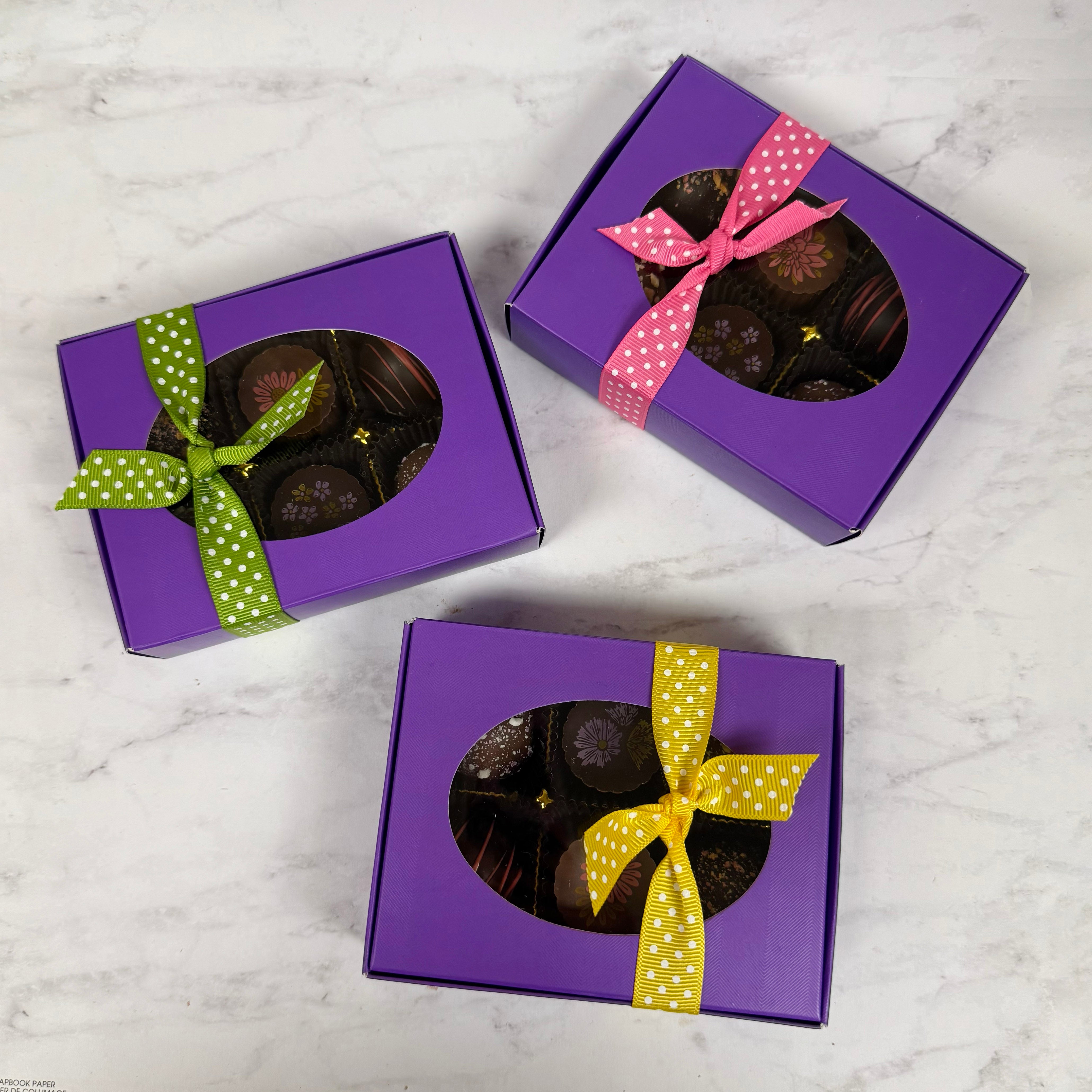 Three purple gift boxes with chocolates and colorful bows on a marble surface.