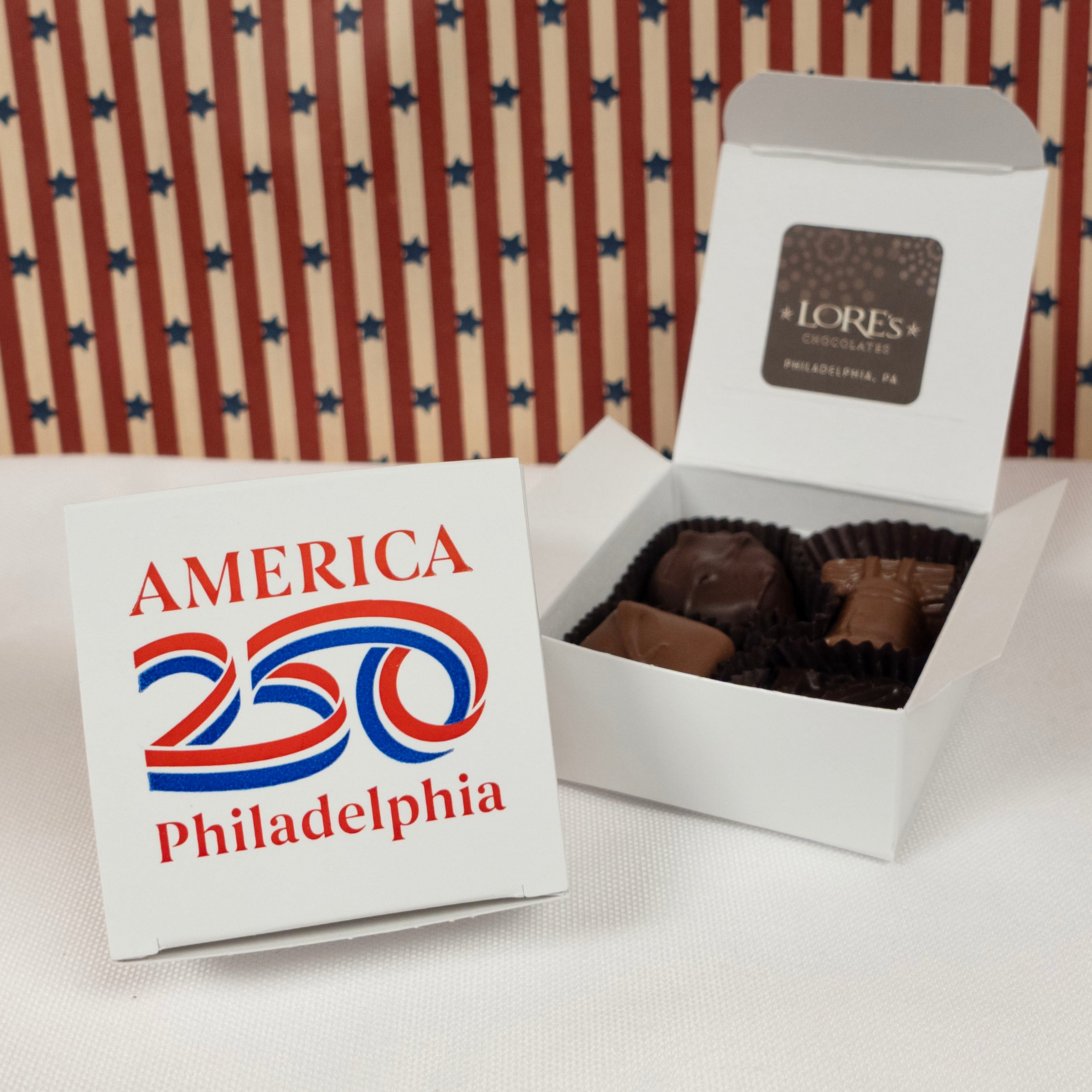 White 4 piece gift box of chocolates with 'America 250 Philadelphia' in red and blue on white surface and a background of stars and stripes. Sticker on inside is blurred but reads 'Lore's Chocolates Philadelphia, PA'