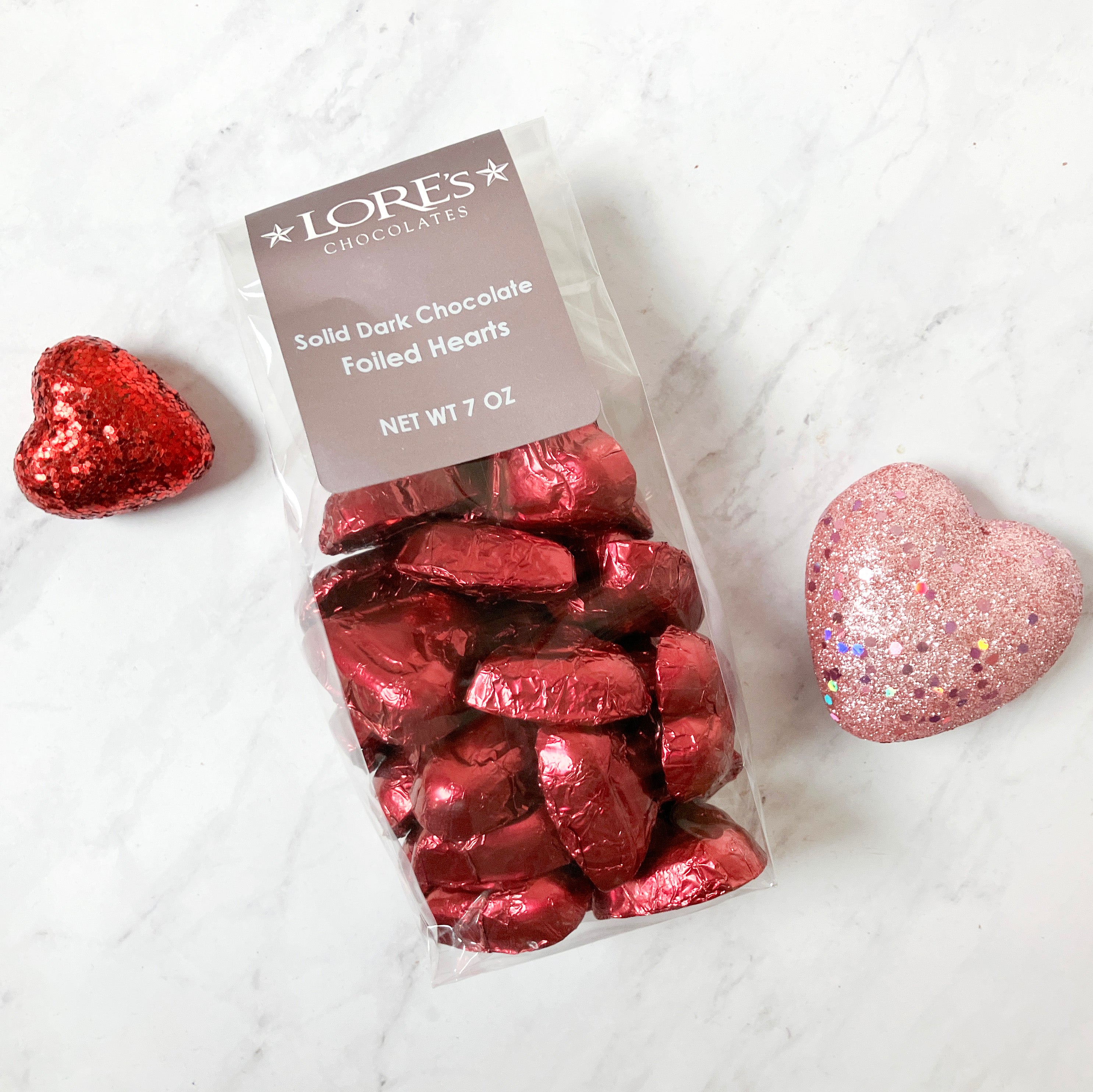 Pack of Lore's Solid Dark Chocolate Foiled Hearts on a white marble surface with two heart-shaped Valentine's decorations.
