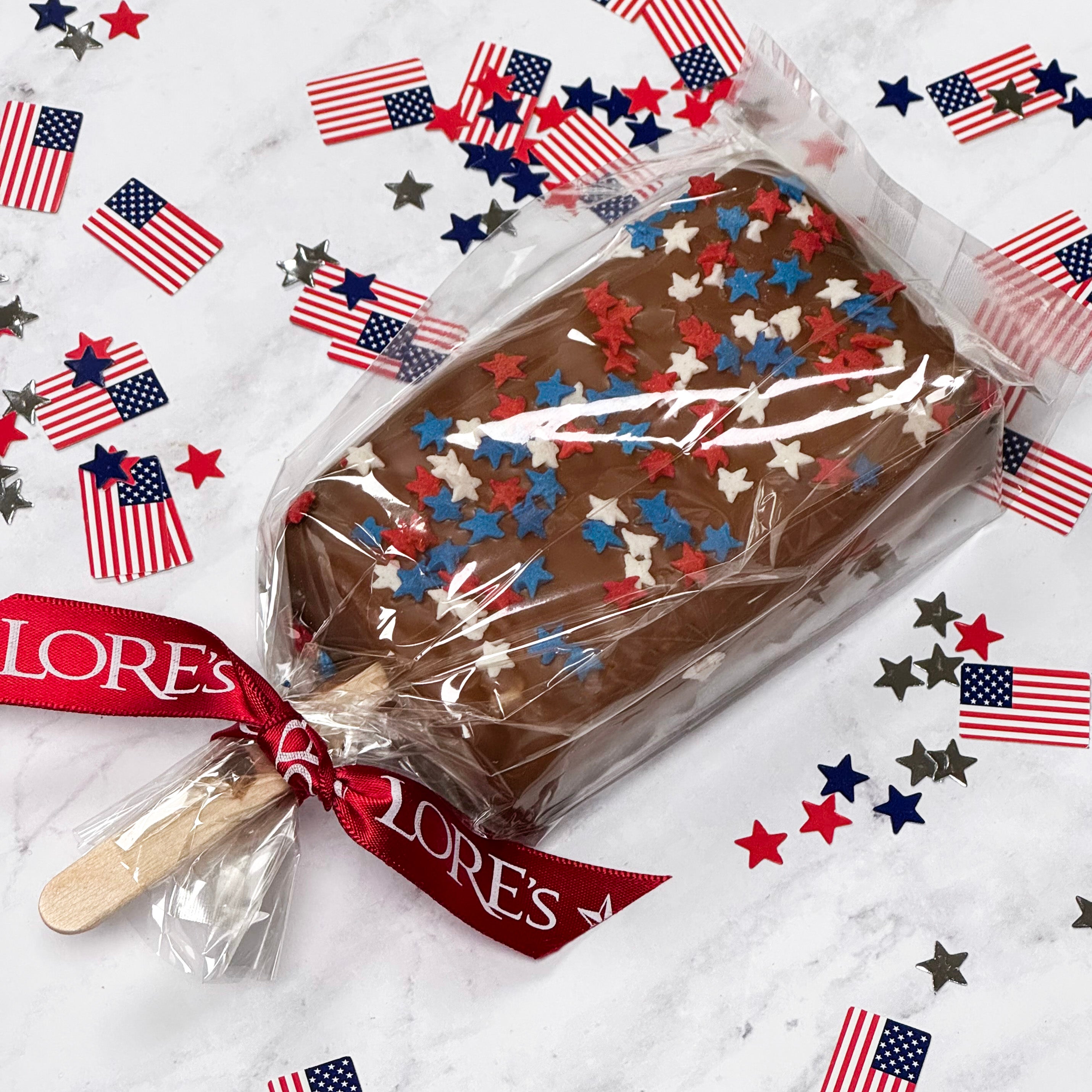 Chocolate-covered Rice Krispies bar with patriotic sprinkles in clear packaging, surrounded by American flags and stars on a white background. Lore's Chocolates in Philadelphia