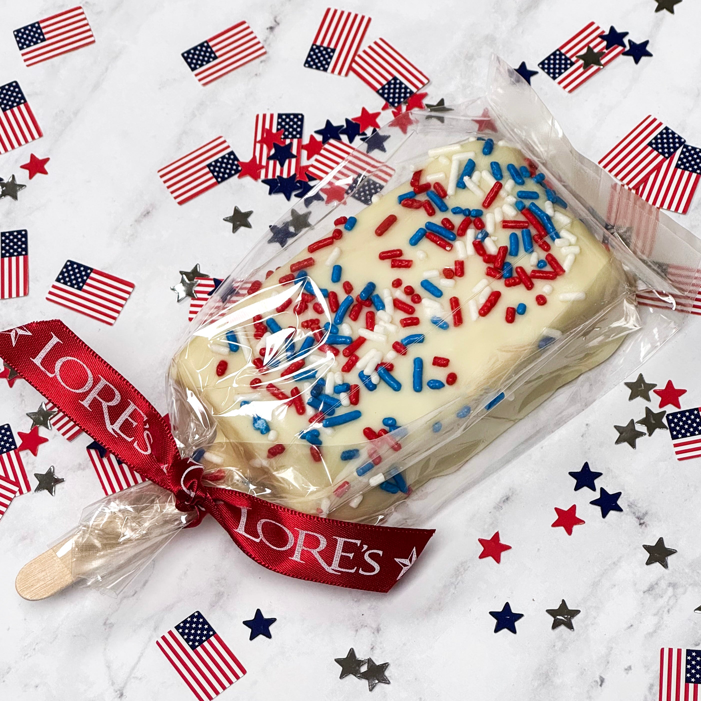 White chocolate-covered Rice Crispies bar with red, white, and blue jimmies / sprinkles on a marble surface with American flag decorations.