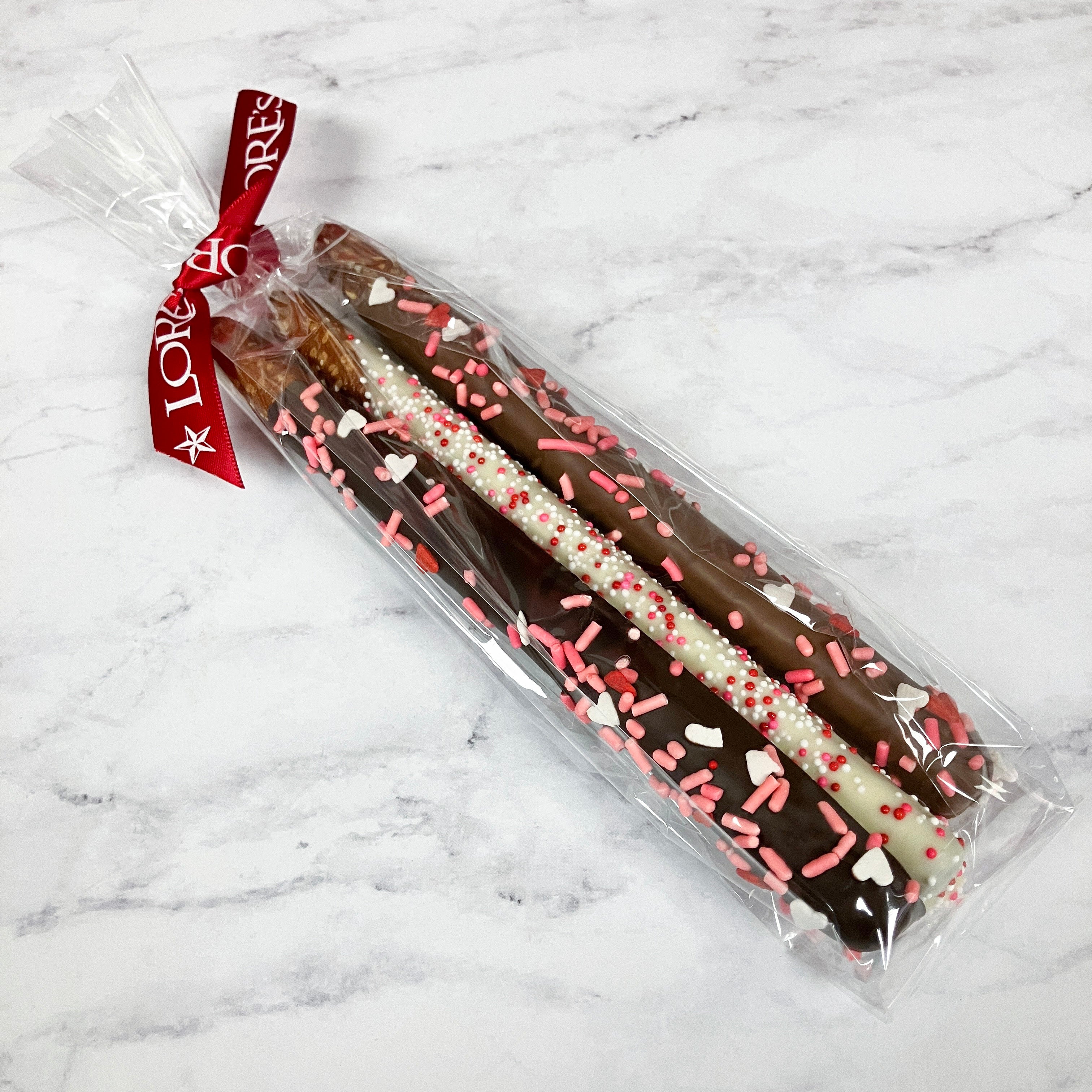Chocolate-dipped pretzel rods with sprinkles in clear packaging on a marble surface