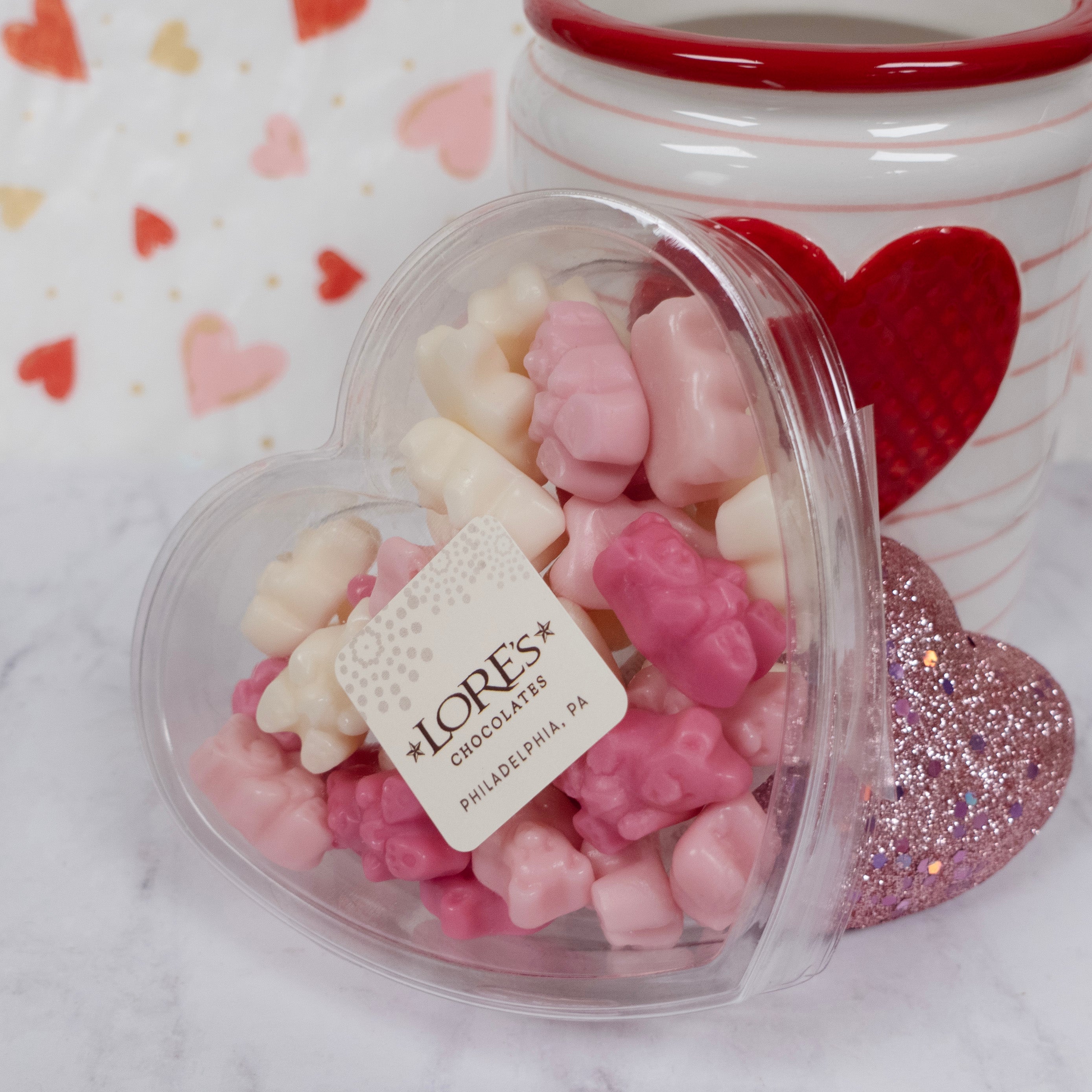 Heart-shaped container of pink and white chocolate pieces with a LORES Chocolates label, on a heart-themed background.