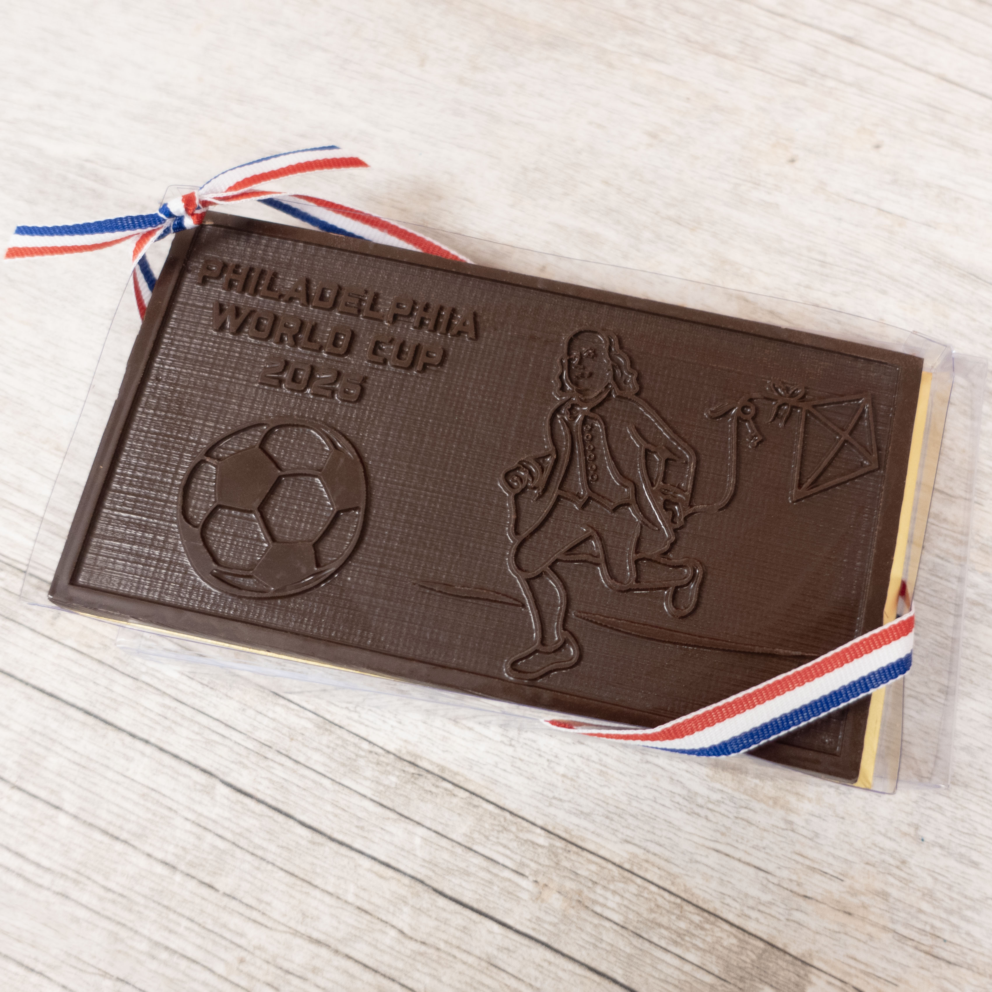 Dark Chocolate bar with Philadelphia World Cup 2026 design and bejamin franklin playing soccer for FIFA on a wooden surface - Lore's Chocolates in Philadelphia