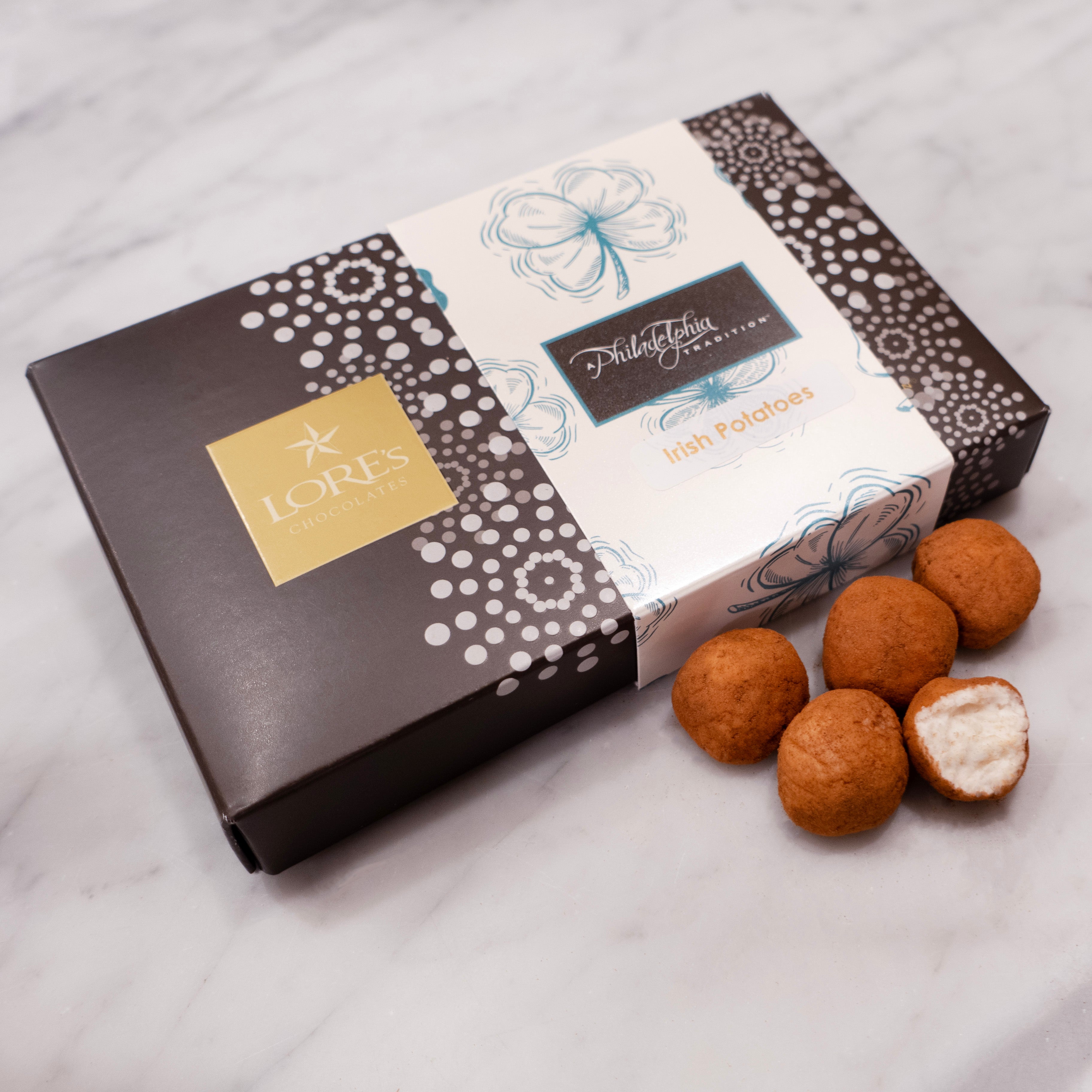half pound box from Lore's Chocolates with a shamrock belly band reading "A Philadelphia Tradition®, Irish Potatoes" Next to the box are 5 cinnamon covered irish potatoes, one open to show the coconut cream inside
