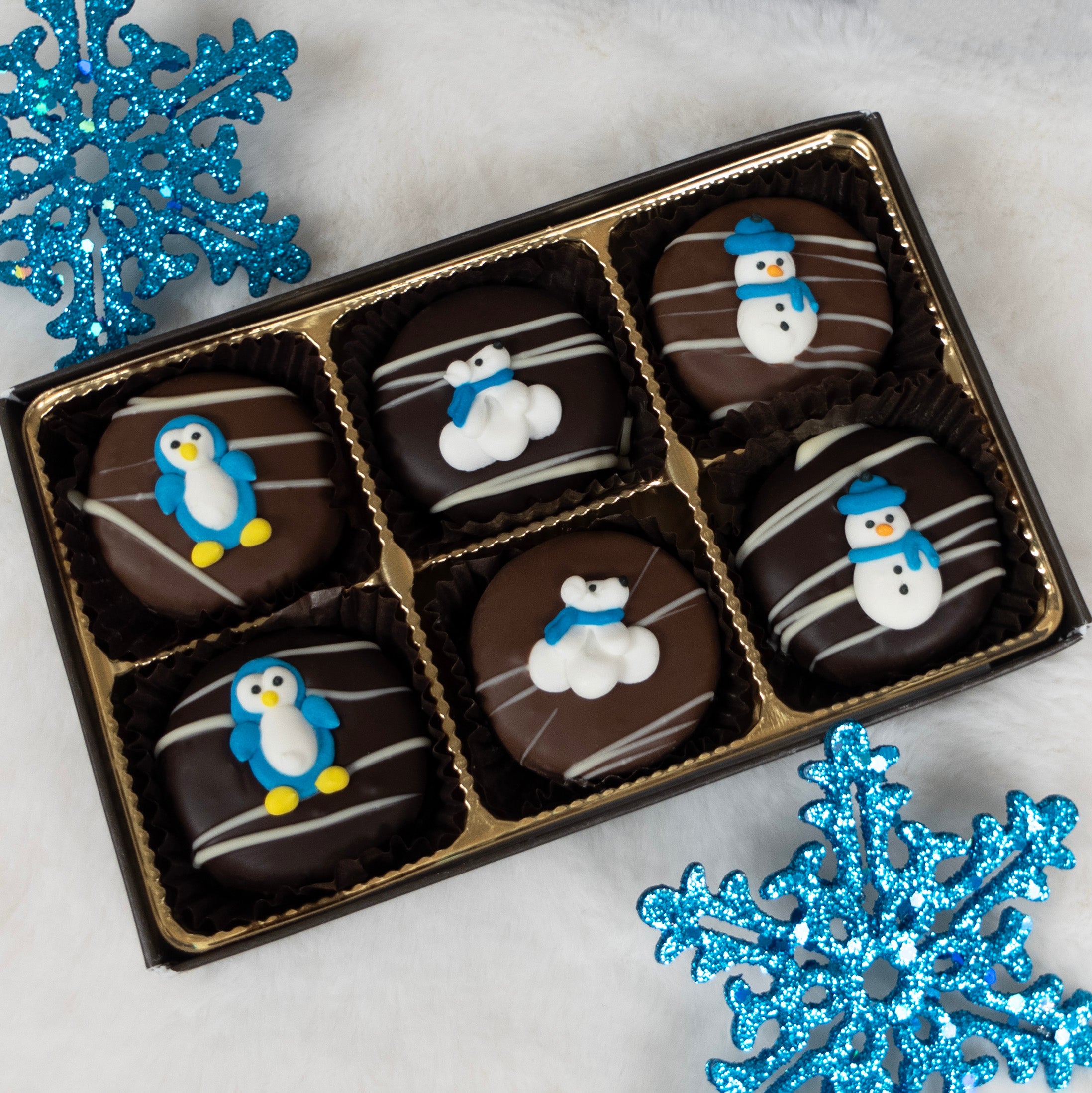 Box of chocolate oreo cookies with snowman, polar bear, and penguin designs, surrounded by blue glittery snowflakes on a light background.
