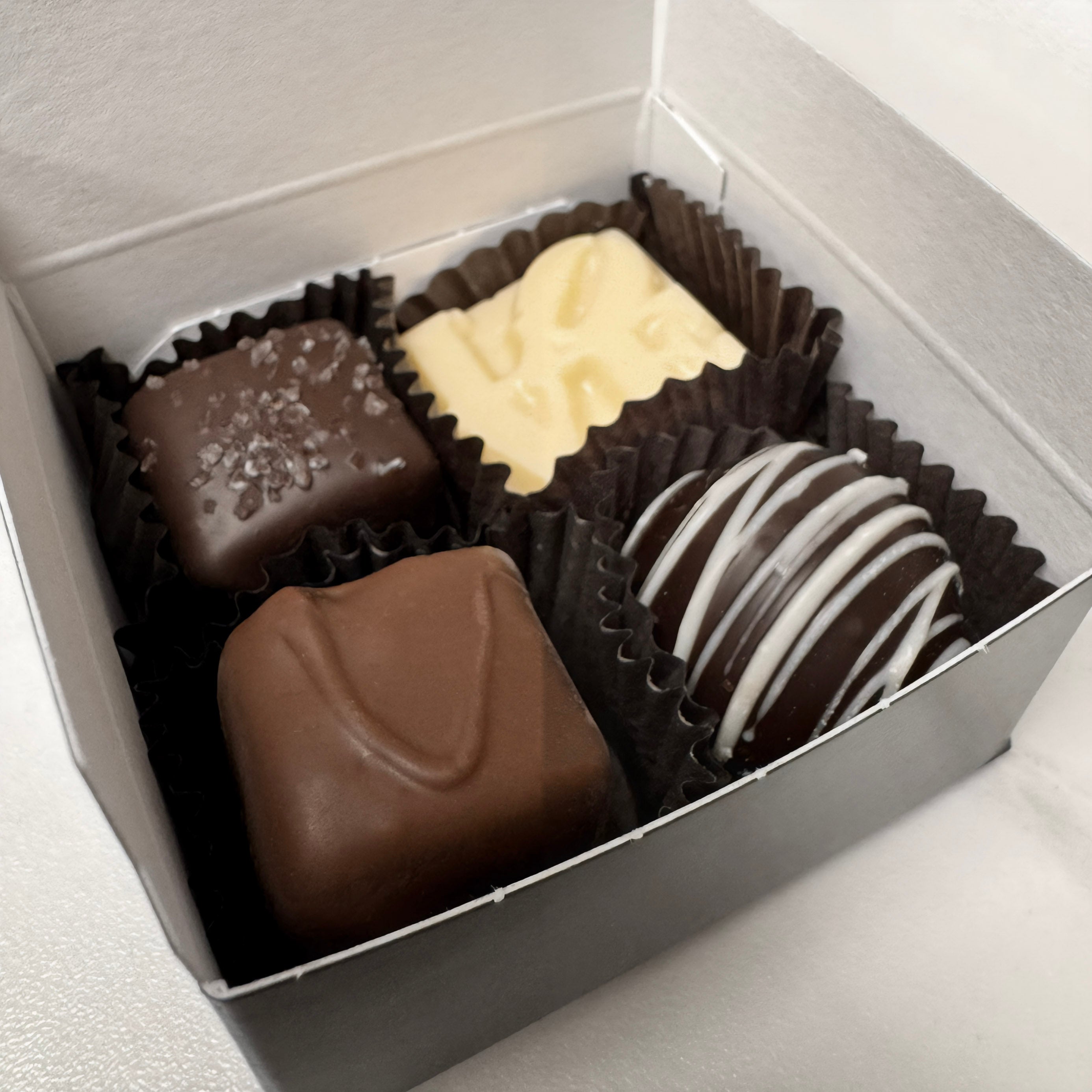 Assorted chocolates close up in a box on a light gray background. includes: dark sea salt caramel, milk vanilla caramel, champagne truffle, and white chocolate LOVE. made by Lore's Chocolates in Philadelphia