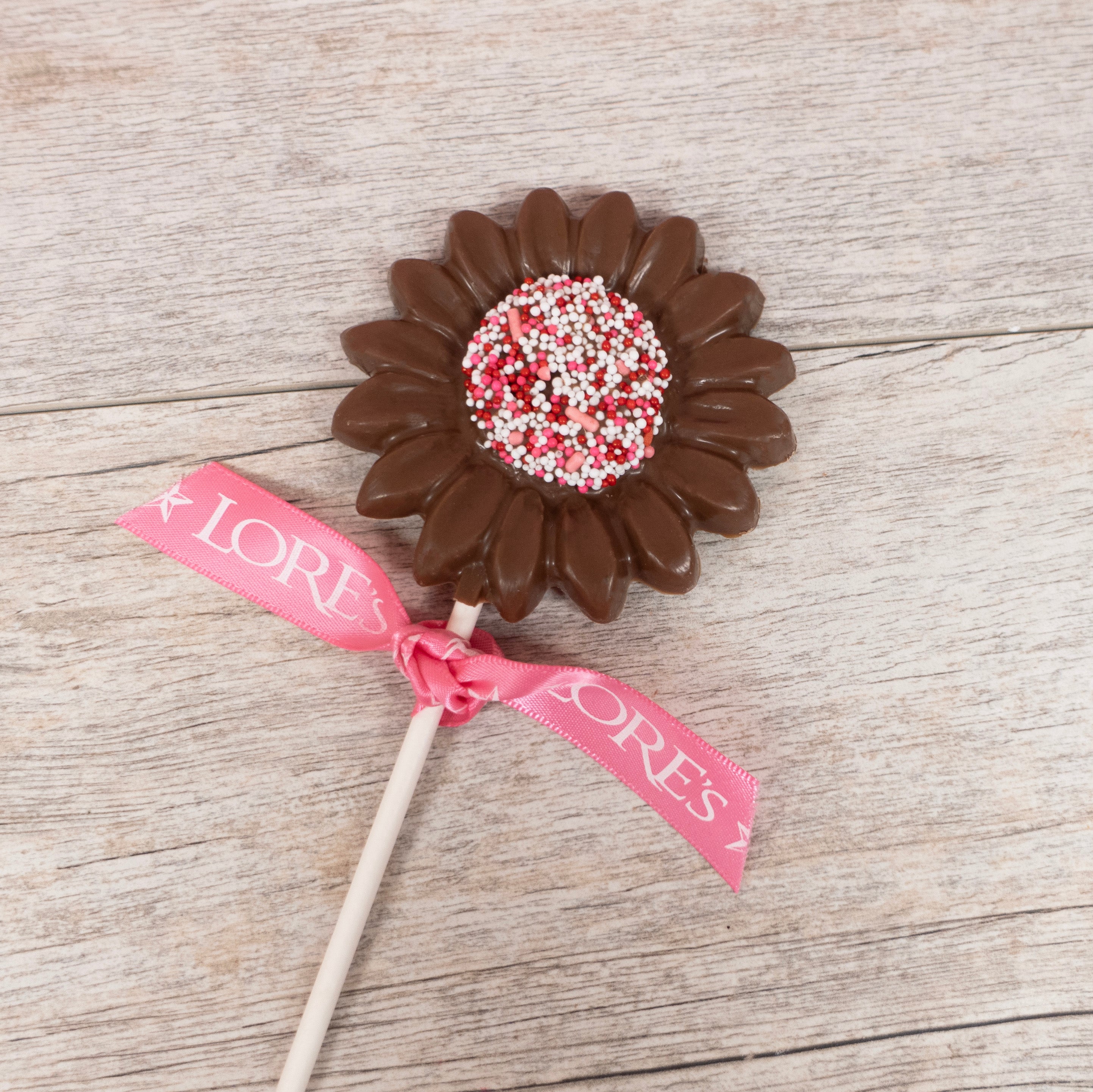 Lore's Chocolate Lollipop in the shape of a sunflower, center is covered in nonpariel seeds in pink, red, and white for valentine's day, pop is tied with a pink Lore's Ribbon