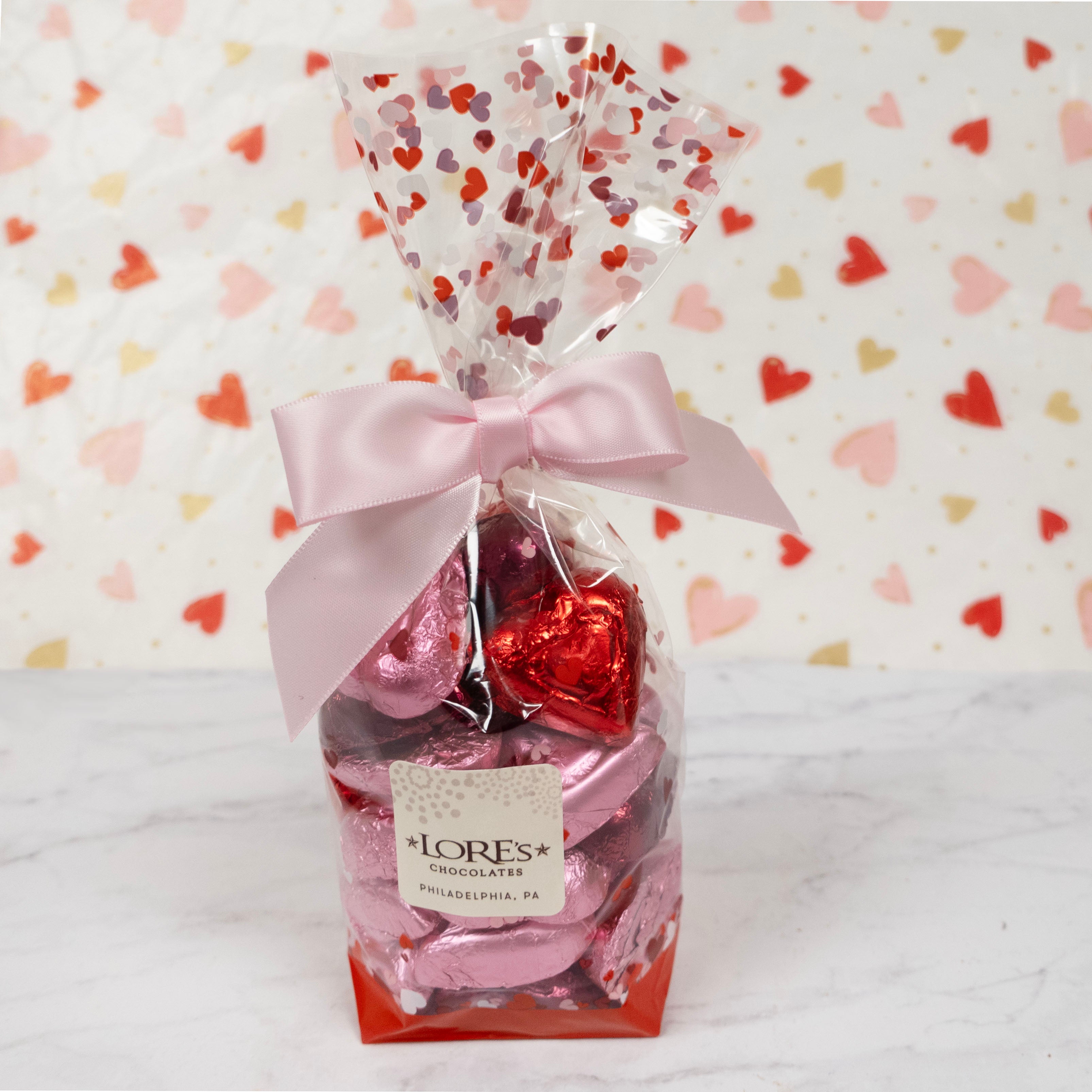 Pack of red and pink heart-shaped foil-covered milk chocolates with LORES Chocolates packaging on a heart-patterned background