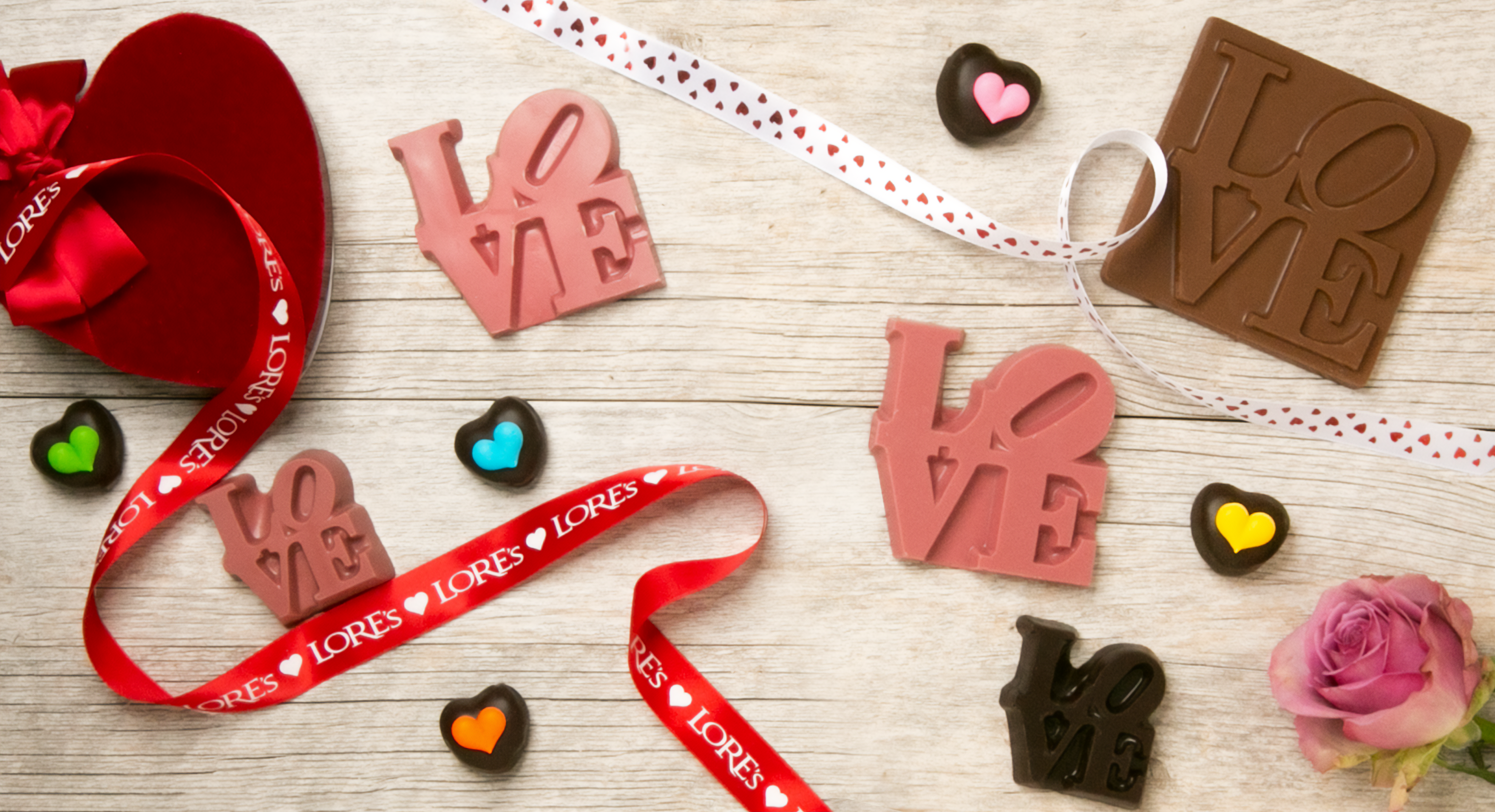 Valentine's Banner with heart-shaped box, ruby chocolate LOVE statues, decorated heart shaped cream filled chocolates, and a pink rose. Along the image is Lore's Ribbons with hearts 