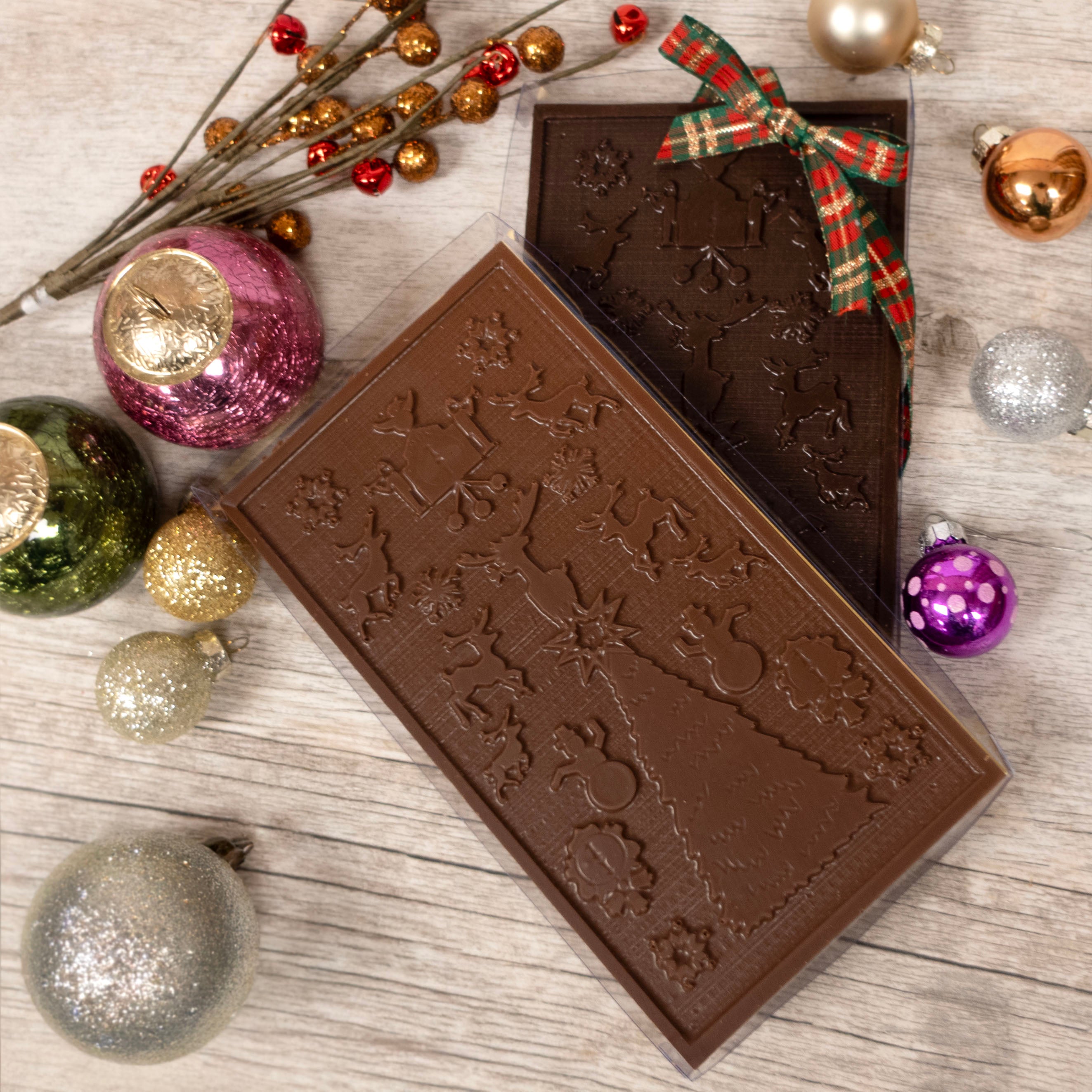 Two chocolate bars with festive designs on a wooden surface with Christmas ornaments.